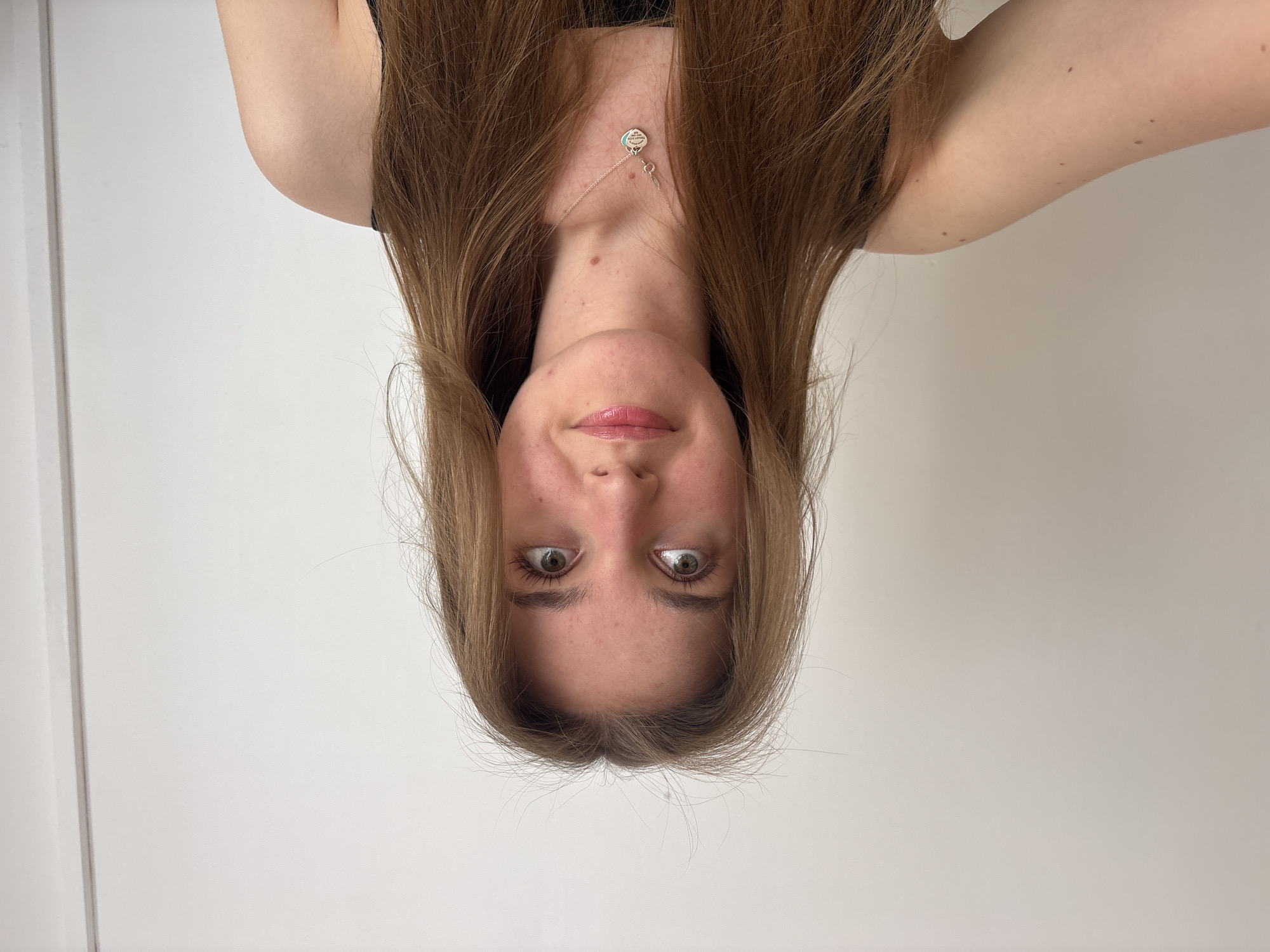 A portrait of a woman with fair skin, light brown hair, and light-colored eyes is presented upside down. She is wearing a dark-colored top and a silver necklace. The background is a plain white wall. The location is not discernible from the image. The image provides no indication of city of Perm, Russia.