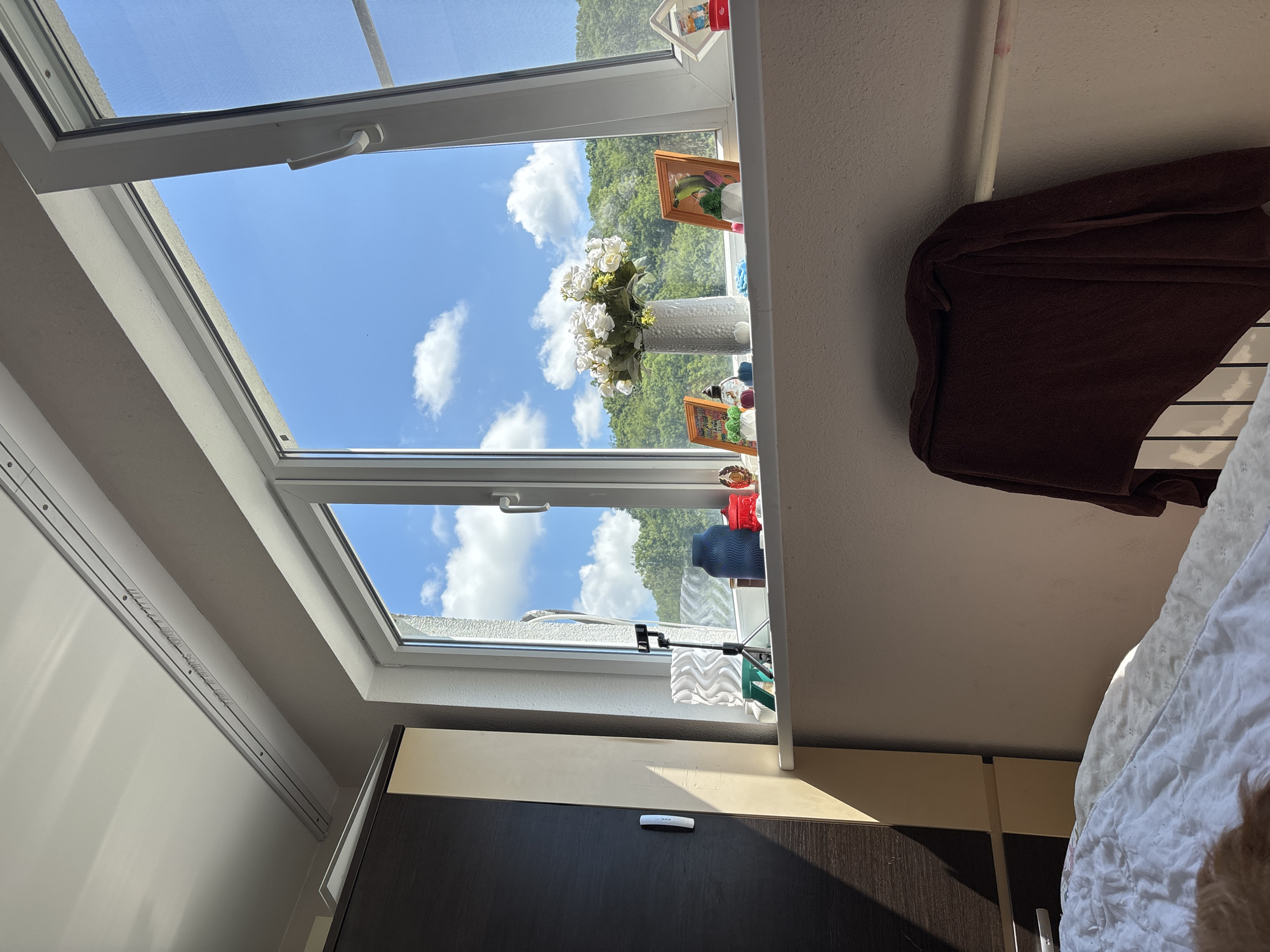 A bedroom scene features a large window overlooking a bright, sunny day with a clear blue sky and scattered white clouds. Lush green trees and hills are visible beyond the window. The windowsill displays several decorative items, including a vase of white flowers, framed pictures, and other trinkets. To the left of the window, a portion of a wardrobe is visible, with sunlight casting a shadow on it. In the foreground, a bed covered with white bedding is partially shown, with a dark brown towel draped over a radiator. The overall impression is that of a peaceful interior setting on a beautiful day, possibly in a residential area of Sochi, Russia, given the typical landscape and architecture seen in the region.