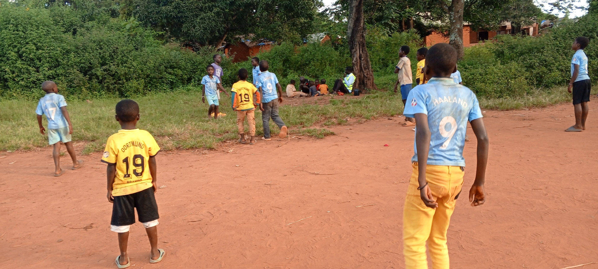 The image depicts a group of African children playing outdoors on a dirt ground in Ngalula, Democratic Republic of the Congo. In the foreground, two boys are seen from the back, one wearing a yellow "Dortmund 19 Brandt" jersey and black shorts, and the other sporting a light blue jersey with "Haaland 9" and yellow pants. Several other children are present in the background, some wearing similar soccer jerseys and others in casual clothing. A group of individuals is seated near a tree in the middle ground. The background features dense green vegetation and simple brick structures. The lighting suggests it is daytime.