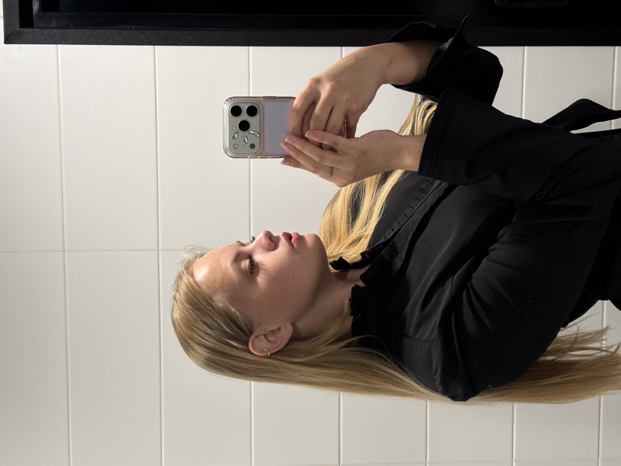 A young blonde woman is taking a mirror selfie in a restroom. She is wearing a black ruffled shirt and is holding a phone with a decorated case. The restroom has white tiled walls. The image appears to have been taken in Izhevsk, Russia.
