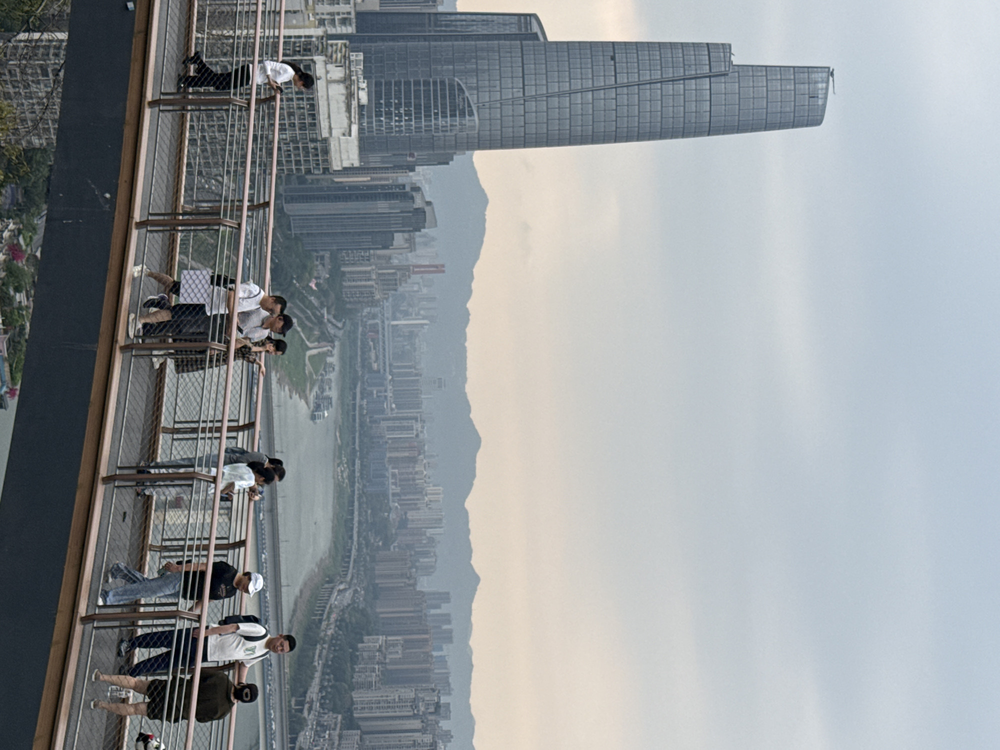 The image depicts a group of people standing on an elevated observation deck in an urban environment. The location appears to be in the Yuzhong District of Chongqing, China, given the distinctive architecture of the Raffles City Chongqing complex, including its horizontal skyscraper, which is visible in the background.
The people are of diverse ages and are dressed in casual attire. They are observing the cityscape below, which includes the Jialing River, numerous buildings, and mountain ranges in the distance. The sky is overcast, providing diffused lighting.
The observation deck is constructed with a metal framework and safety barriers. The composition of the image suggests a high vantage point, giving a comprehensive view of the city.