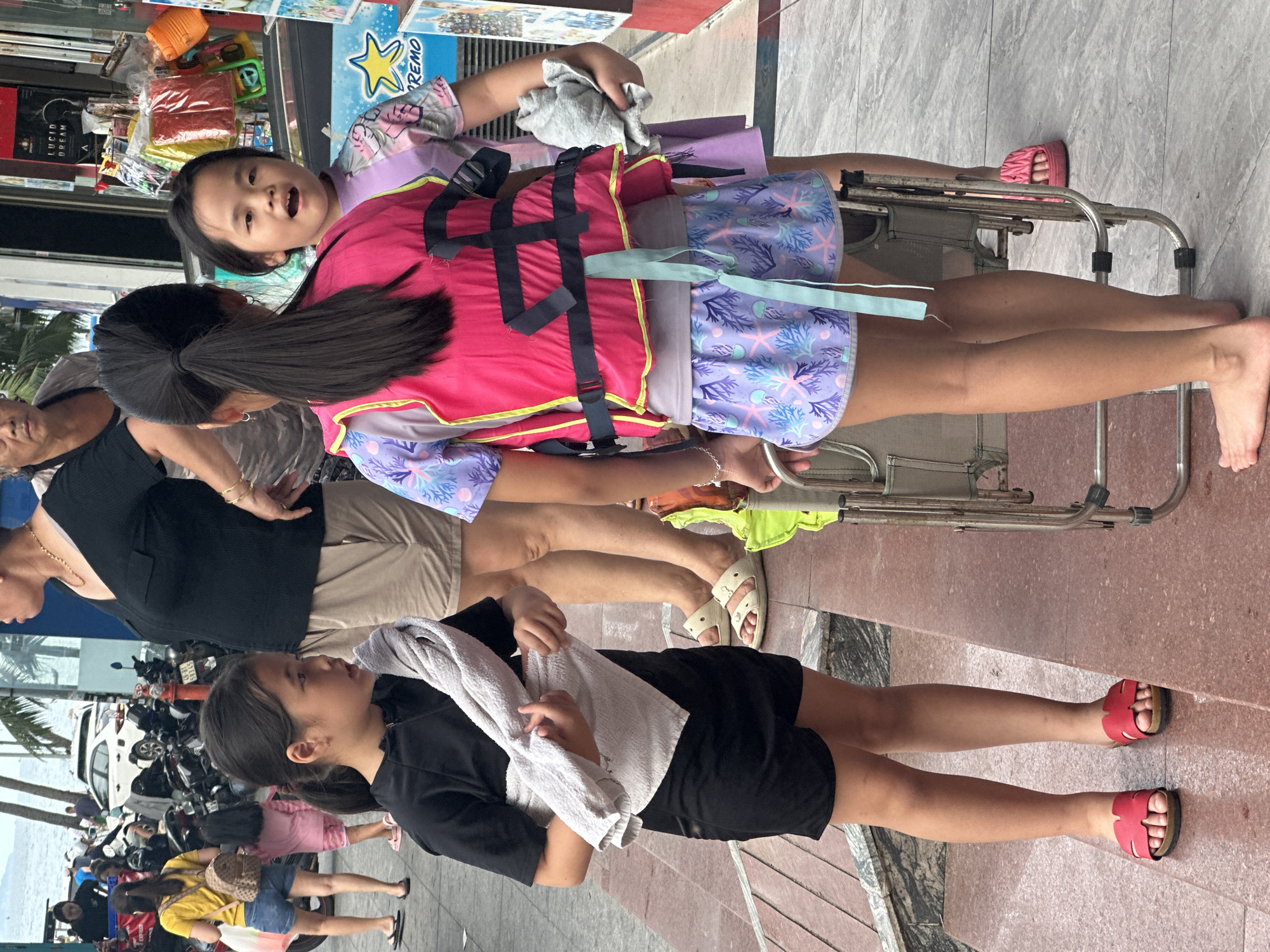 The image depicts a scene in Bac Nha Trang Ward, Vietnam. A young girl in a black outfit holds a white towel, standing on a sidewalk lined with red pavers. A second girl, wearing a pink life vest over a lilac and blue swimsuit, sits on a metal-framed transport chair. An older woman in a black top and khaki shorts stands nearby. In the background, a street scene includes parked motorcycles, a white car, palm trees, and the ocean. A variety of shops and merchandise are visible along the sidewalk. Pedestrians walk in the background.