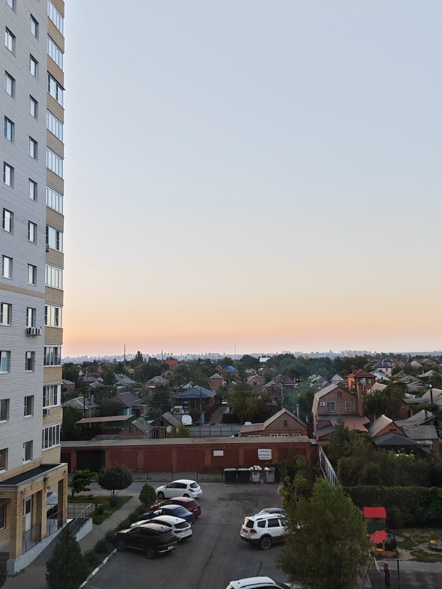 The image shows a view of the city of Bataysk, Russia, taken from an upper floor of a residential building. The building itself, with its light-colored siding and numerous windows, occupies the left side of the frame. Below the building, a parking lot is visible with several cars parked, including a white SUV, a dark SUV, a white hatchback, a blue sedan, and a red hatchback.
Beyond the parking lot, the scene transitions to a residential neighborhood characterized by a dense collection of houses with various roof styles and colors. Green trees and foliage are scattered throughout the neighborhood, suggesting a temperate climate. In the background, a cityscape with taller buildings is visible on the horizon, silhouetted against a pale orange and blue gradient sky, indicative of either sunrise or sunset. There are no people clearly visible in the image. The overall impression is a suburban or semi-urban residential area within the city.