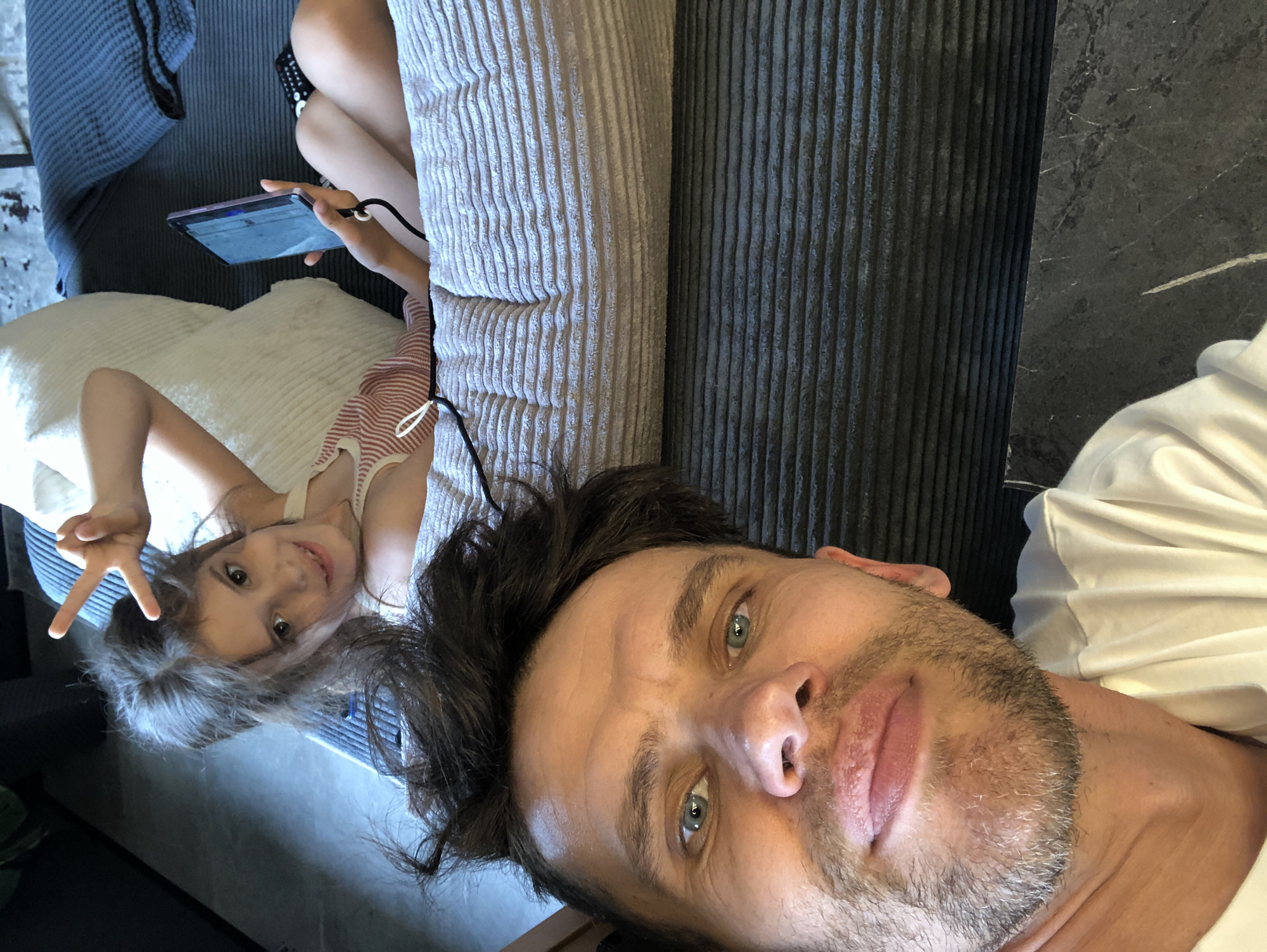In an indoor setting, a man and a young girl are present. The man is in the foreground, looking towards the camera with a slight smile, his face close to the lens. He has short, dark, messy hair and blue eyes, with a light beard and stubble. He is wearing a white t-shirt.
Behind him, a young girl lies on a textured, light-colored surface, possibly a sofa or bed, with a large, white, textured cushion behind her. She is looking at the camera and smiling, with her right hand raised in a peace sign gesture. She has long, dark brown hair that is partially covering her face. She is wearing a striped tank top. To her right, she is holding a tablet device in her left hand. A remote control is visible near her legs.
The environment suggests a domestic indoor space, likely a living area. The flooring appears to be dark and textured, with horizontal lines. The background includes dark blue fabric, possibly blankets or upholstery. There are no discernible external city landmarks or indications of Saint Petersburg, Russia within the image itself.