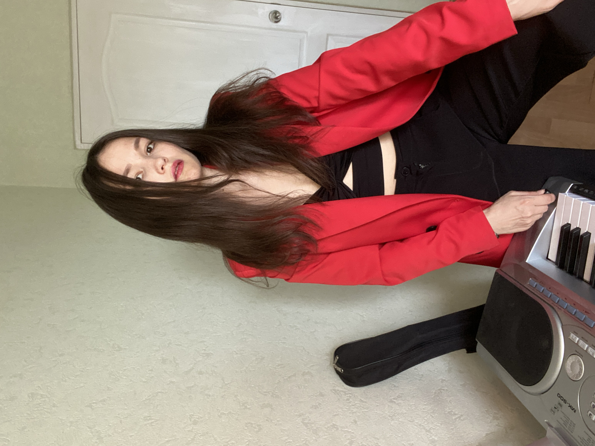 A young adult female with long, dark hair is posing in an indoor setting. She is wearing a bright red blazer over a black crop top and black pants. To her left, a black case rests against a textured, pale green wall. In front of the case, a silver and black electronic keyboard is visible, with its keys facing the viewer. A closed white door with a round doorknob is situated to the right of the woman. The scene is lit with soft, natural light. The location appears to be an apartment in the city of Chaikovsky, Russia, given the typical interior design elements and the absence of any outdoor elements.