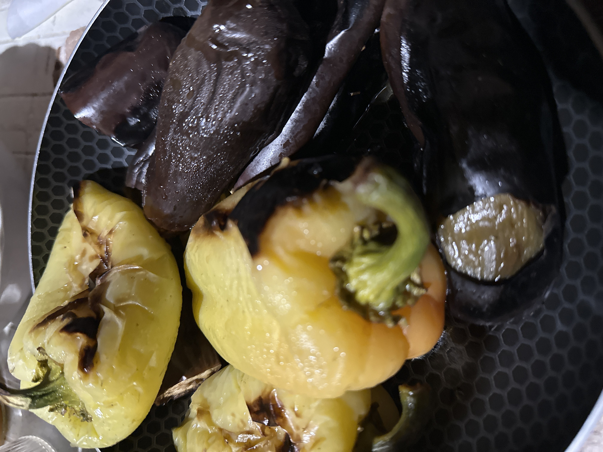 The image shows a close-up, overhead view of a non-stick pan containing cooked yellow bell peppers and dark purple eggplants. The vegetables appear to be roasted or stewed, with some areas of charring on the peppers. The pan has a honeycomb-like pattern on its surface. The setting is indoors, with a light-colored surface visible in the background. No specific location in Sochi, Russia can be determined from the image. There are no people, animals, or any other identifiable objects or actions present.
