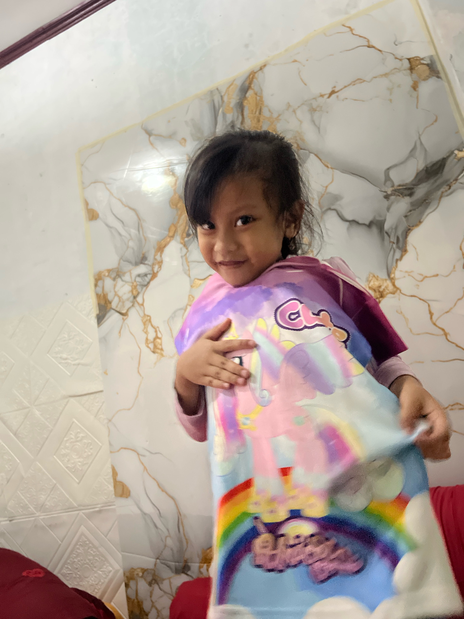 A young girl with dark hair, wearing a pink and purple long-sleeved shirt and a t-shirt featuring a unicorn and rainbow graphic, smiles at the camera. She is holding the t-shirt open with her hands. The background consists of a wall with a marble pattern and a textured white section. The scene appears to be indoors, possibly in a residential setting in Kecamatan Pati, Indonesia.