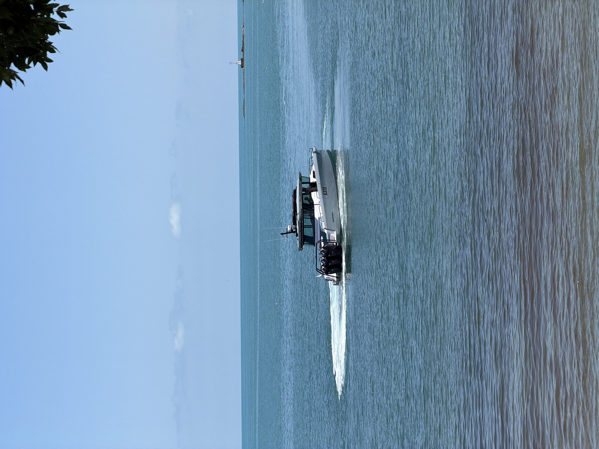 A white motorboat with the name "KAYA" on its side is traveling through turquoise waters, creating a white wake. The boat has a dark cabin structure on top and triple outboard motors at the stern. In the distance, a small lighthouse stands on a rocky outcrop. The sky is a clear light blue with a few wispy clouds. The foreground shows calm, rippling water with a sandy bottom visible beneath the surface. The image appears to have been taken from a shore or a slightly elevated position, with some dark green leaves hanging from the top right corner. The specific location in Tsarevo, Bulgaria is not identifiable from the visual information provided.
