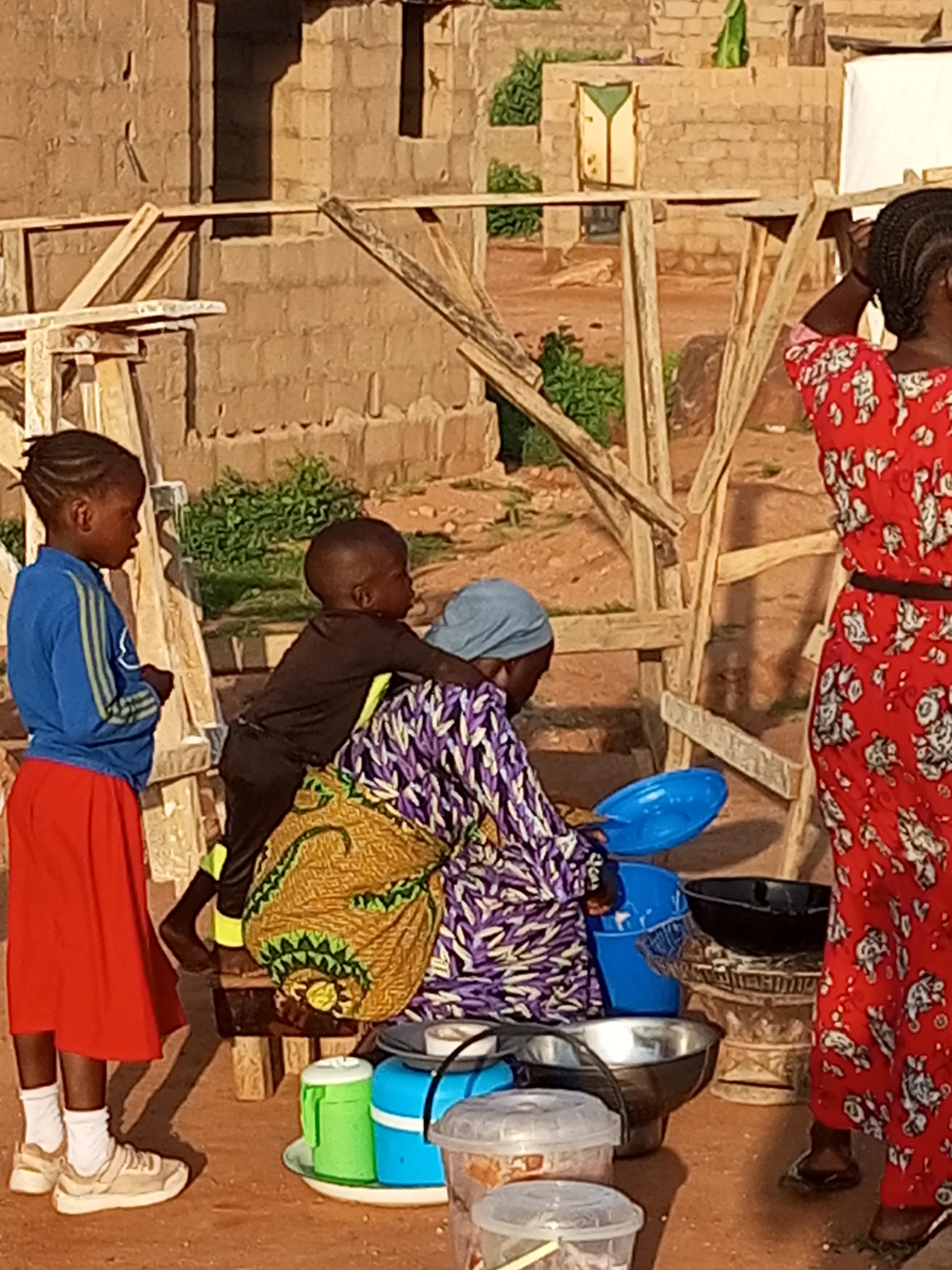 In Pantame, Nigeria, a scene unfolds involving four individuals and various objects near a partially constructed building. A young girl on the left, wearing a blue long-sleeved shirt with yellow stripes and a red skirt, stands looking towards the right. Next to her, a young boy in a black long-sleeved shirt and black pants leans on a woman. The woman, wearing a purple and white patterned top and a light blue headscarf, is holding a blue plastic basin and appears to be interacting with the boy. Her back is to the camera, and a patterned cloth bag with green and yellow designs is draped over her shoulder. To the right, a woman in a red dress with white floral patterns stands with her back to the camera, adjusting her hair. Several objects are present on the ground, including a large metal basin, a blue plastic bucket, a black bowl, a green and white thermos, a blue container, and two transparent plastic containers with lids. The ground is reddish-brown dirt, and wooden scaffolding structures are visible throughout the scene. The background shows a brick building under construction with openings that suggest windows or doorways.