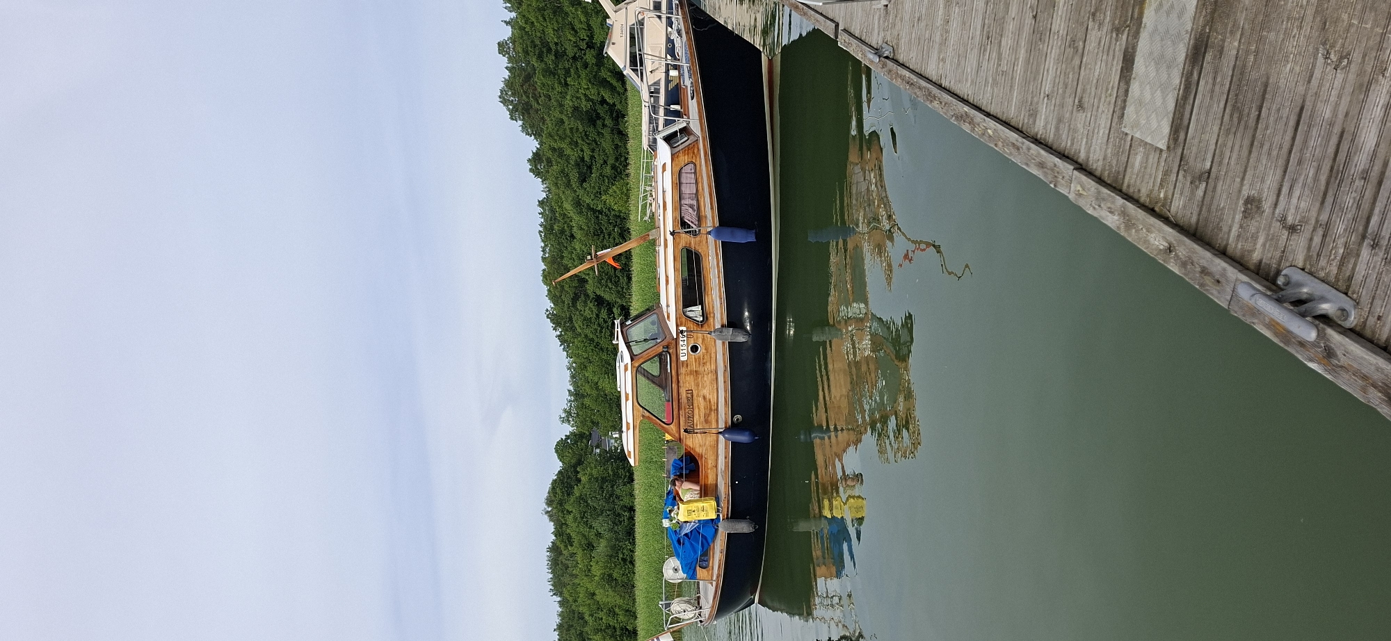 A motorboat with a dark blue hull and wooden superstructure is moored alongside a wooden dock. The boat is gently rocking on calm, green water, creating a clear reflection. The boat has a covered cabin with windows and a small deck area. Two blue fenders are visible along the hull. A person is partially visible inside the cabin, reclining on a seat. The background features lush green trees and a slightly overcast sky. The wooden dock is weathered and shows signs of use, with metal cleats attached to it. The scene appears to be a marina or a calm waterway, consistent with locations in Naantali, Finland, known for its archipelago and boating activities.