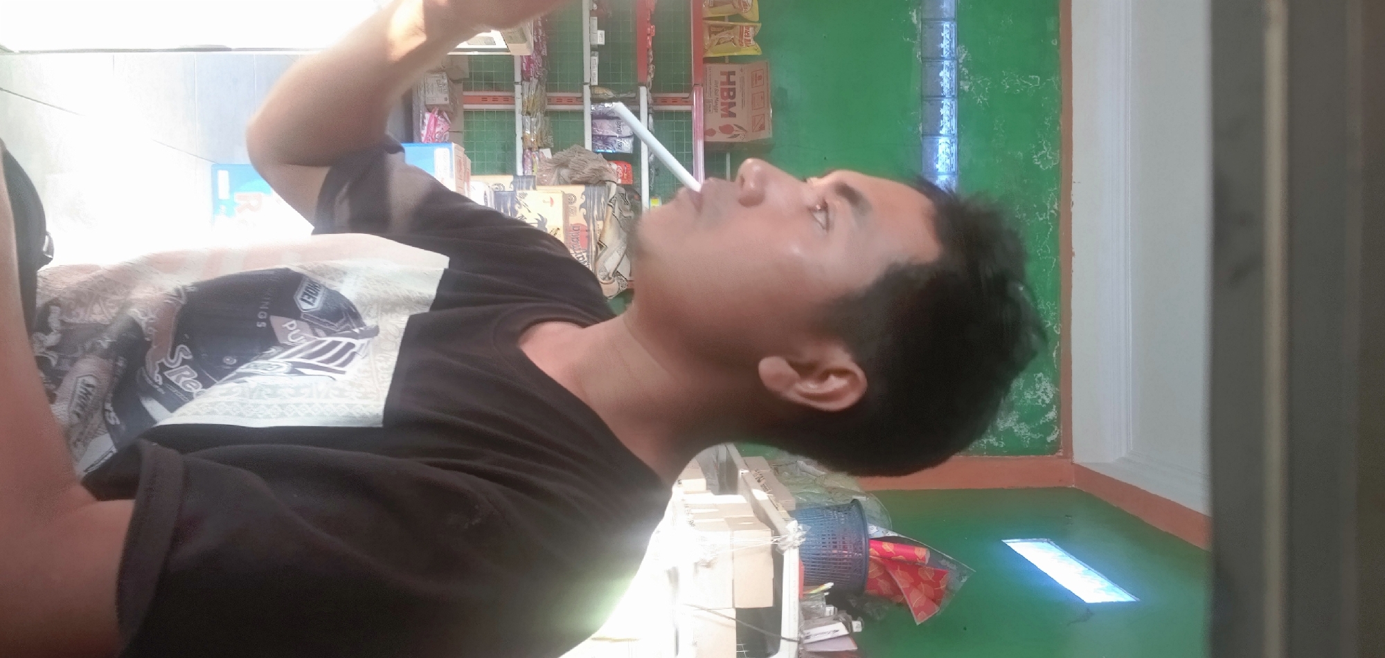 A man is pictured in profile, looking to the left and smoking a cigarette. He is wearing a black t-shirt with a graphic design on the front. The background appears to be a room or store, with shelves stocked with various items, including boxes and what look like snacks or food products. The walls are painted green, and there is a window with light coming through. The overall setting suggests an indoor environment, possibly a small shop or a home. The man's posture and the presence of a cigarette indicate a casual moment.