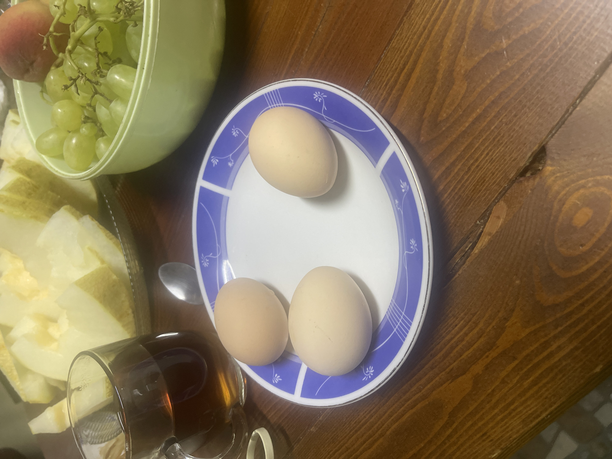 On a dark brown wooden table, a light blue and white plate holds three light brown eggs. To the left of the plate, a clear glass mug is filled with dark brown tea. Behind the mug, sliced pieces of light yellow melon are arranged on a light-colored plate or platter. To the right of the plate of eggs is a light green bowl containing a bunch of green grapes and a peach. The scene appears to be set indoors, possibly in the city of Choktal, Kyrgyzstan, though this location is not explicitly depicted in the image.