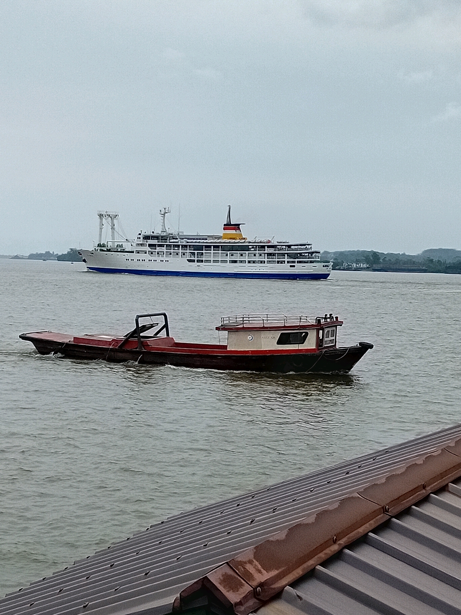 A long, white passenger ship with multiple decks and a red and yellow funnel sails on the water in the background. In the foreground, a smaller, dark-hulled boat with a red deck and a cabin is moving through the water. The boats are on a river under a cloudy sky. The foreground also shows a dark, corrugated metal roof. The scene appears to be in the city of Sungai Meriam, Indonesia.