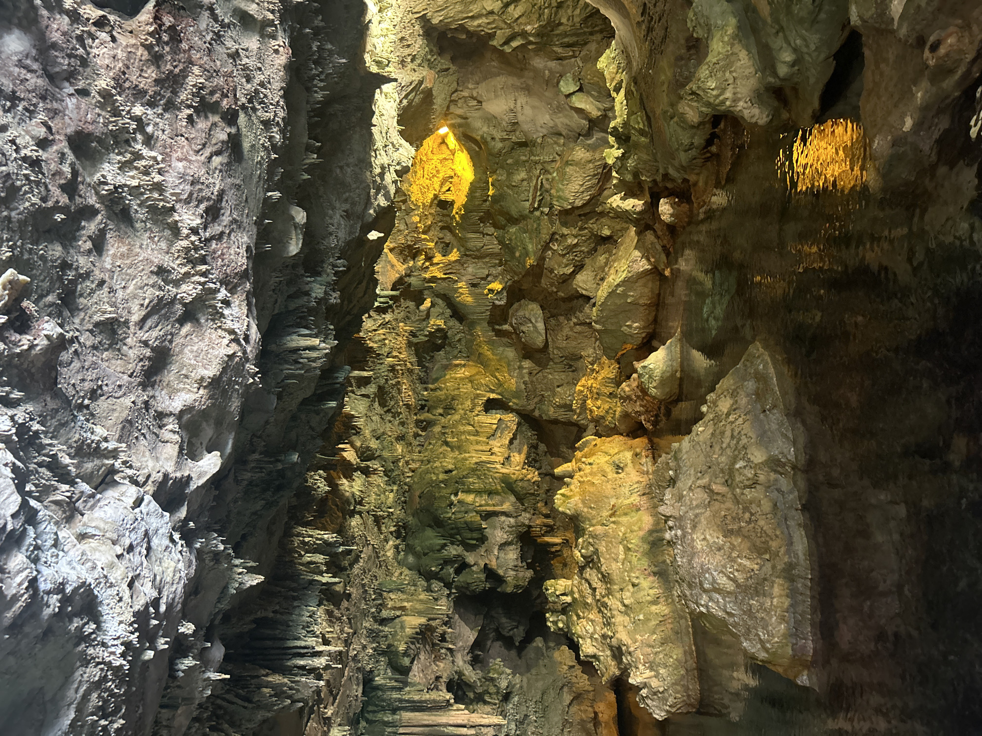 The image shows the Grotta di Nettuno (Neptune's Grotto) in Alghero, Sardinia, Italy. The interior of the cave is characterized by rough rock formations, stalactites, and stalagmites. Some parts of the cave are illuminated with artificial yellow-toned lighting, while other areas are shrouded in shadow. Water is present, reflecting the illuminated areas. The rock surfaces are various shades of grey and brown, with some greenish hues. The cave's architecture is primarily natural, with no immediately visible signs of human alterations other than the lighting.
