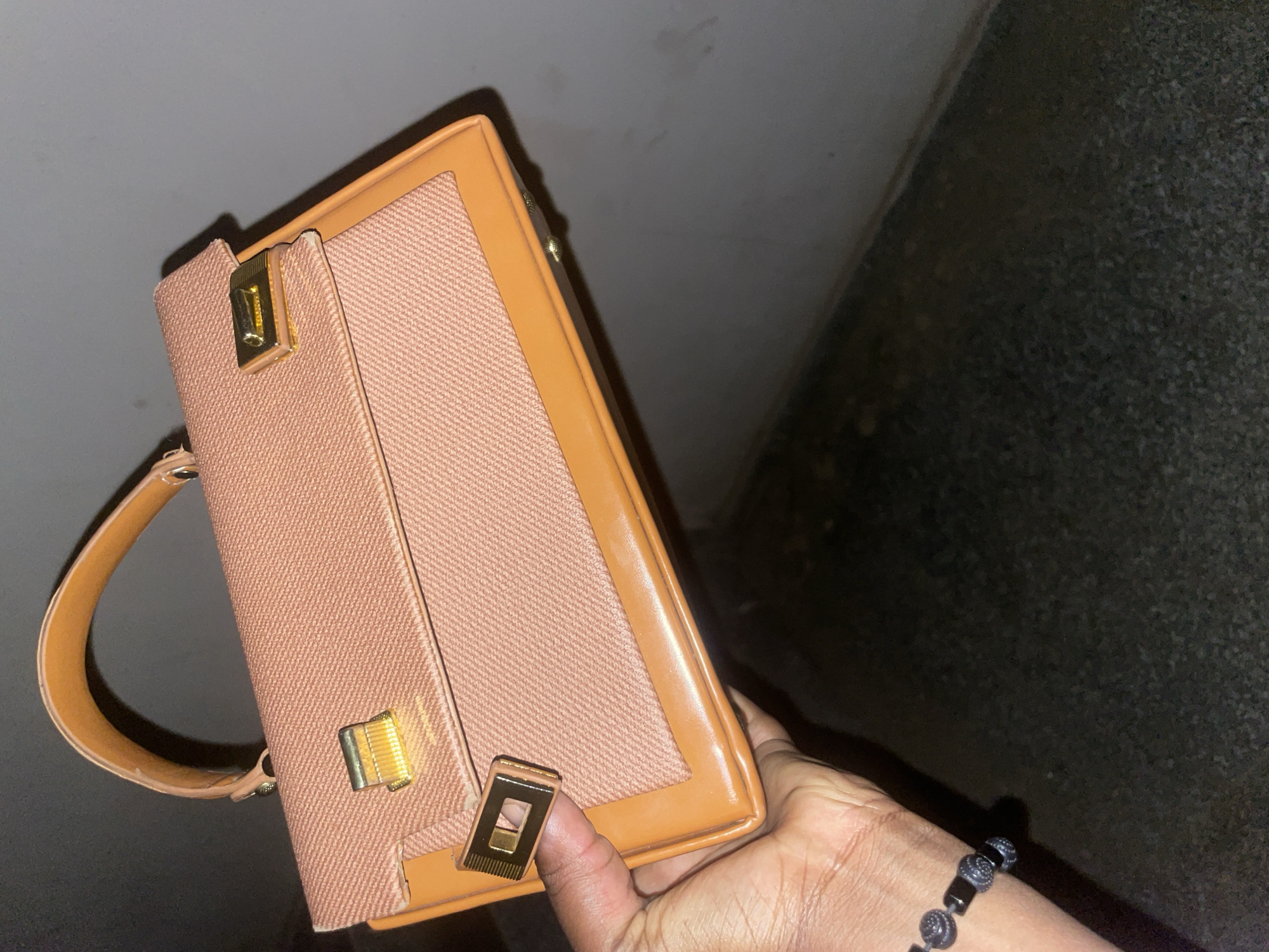 A close-up, angled shot features a structured handbag held by a person's hand against a dark, indistinct background. The handbag is primarily a light peach or pinkish-brown color, made from a textured fabric, possibly canvas. Its edges and a prominent curved handle are crafted from a smooth, light caramel-colored material, likely leather or faux leather.
The bag is adorned with gold-toned metal hardware. A clasp or decorative element near the top of the front flap bears a faint, partially legible inscription that appears to read "HERMÈS". A second, rectangular gold-toned clasp with a central cut-out is visible on the lower part of the flap, positioned near the thumb of the hand.
The hand holding the bag belongs to a person with a dark skin tone. A black beaded bracelet, composed of both textured round and smooth rectangular beads, is worn on the wrist. The lighting is dim, with the handbag well-illuminated in the foreground, suggesting an indoor setting or low-light conditions. The background remains out of focus and very dark, providing no specific location details.