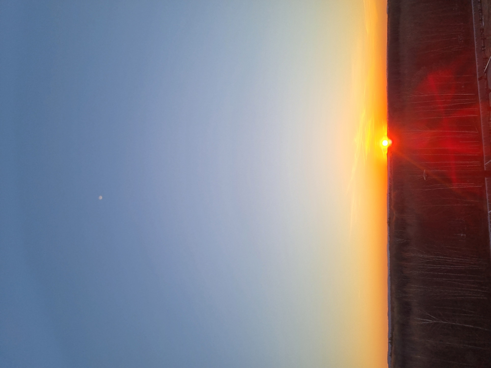 The image shows a sunset. The sun is visible as a bright orange orb on the horizon. The sky transitions from a clear blue on the left to orange near the sun, and then to a darker red on the right. On the left, a small moon is visible in the blue sky. The horizon is obscured by what appears to be a dark field with faint white lines or streaks across it. A blurry fence-like structure is visible on the right edge of the image. It is implied that this image was taken in Kemul, Russia.