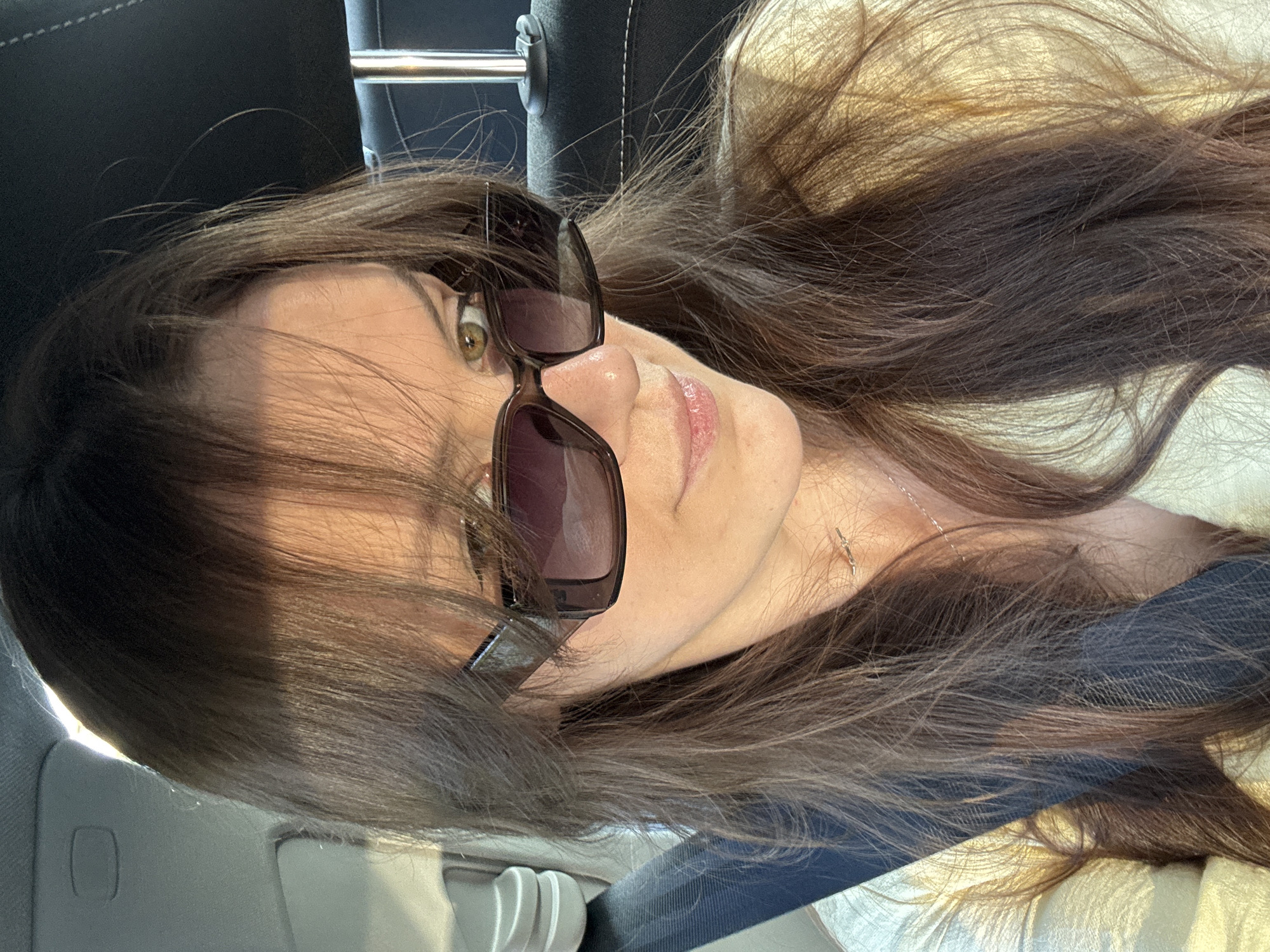This is a close-up, low-angle, portrait-oriented photo of a woman. She has long, dark brown hair that is falling around her face and shoulders. She is wearing large, dark sunglasses that cover part of her eyes. Her skin is fair, and she has on a light-colored shirt or jacket. A sliver of a necklace is visible on her neck.
The lighting suggests it is daytime, possibly late afternoon given the warm tones and shadows. The background appears to be the interior of a car, with a dark seat visible on the left and a lighter, textured surface on the right. There are some subtle metallic reflections, perhaps from a car window or trim. The overall mood is casual and relaxed, with the woman possibly taking a selfie while in a vehicle. No visible text is present in the image.
