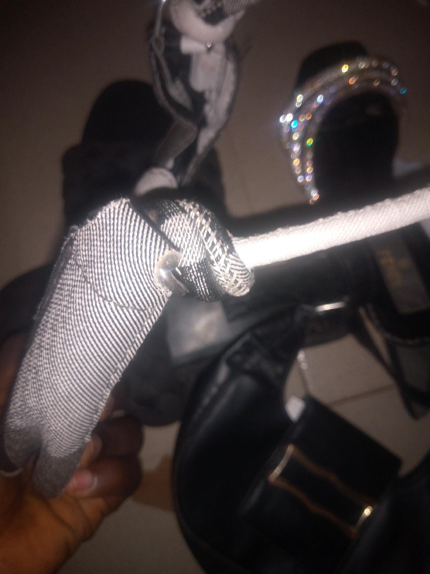 The image shows a close-up of a black handbag with a white rope and a decorative headband with rhinestones. The handbag has a shiny buckle detail. A person's hand is visible holding the rope. The background is dark and out of focus, making it difficult to determine the exact location or environment. There are no discernible landmarks or indicators to suggest it is located in Jalingo, Nigeria. The lighting is dim, suggesting it might be indoors or at night. The image quality is poor, with significant blur and noise.