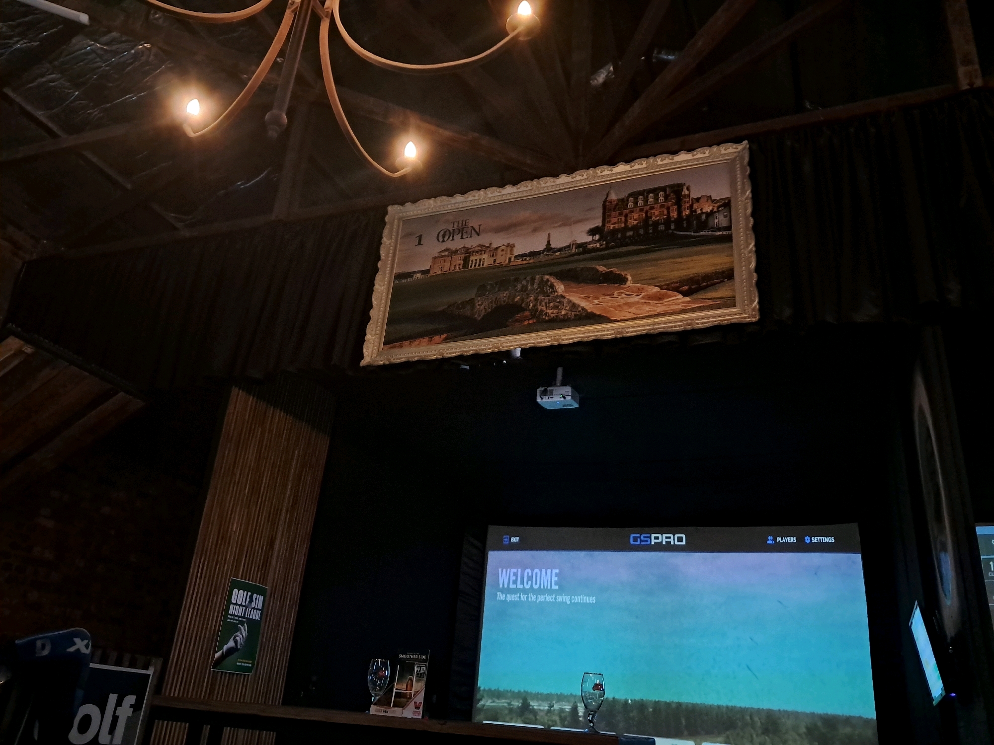 An interior scene captured in the city of Potchefstroom, South Africa, shows a dimly lit room that appears to be a recreational facility, possibly a sports bar or entertainment venue. A large, framed painting of a historical building and a golf course, titled "The Open," hangs prominently on a dark wall, illuminated by a vintage-style chandelier with four bulbs. Below the painting, a large screen displays a golf simulator interface, with the words "WELCOME" and "The quest for the perfect swing continues" visible. The screen also shows the brand "GSPRO" at the top. On the shelf in front of the screen are two wine glasses, one containing a red liquid, and a small brochure. To the left, a poster for "GOLF SIM NIGHT LEAGUE" is attached to a slatted wooden wall. A blue chair with a white logo is partially visible in the lower left corner, along with another sign that reads "olf." The overall ambiance is dark and moody, with lighting focused on the painting and the screen.