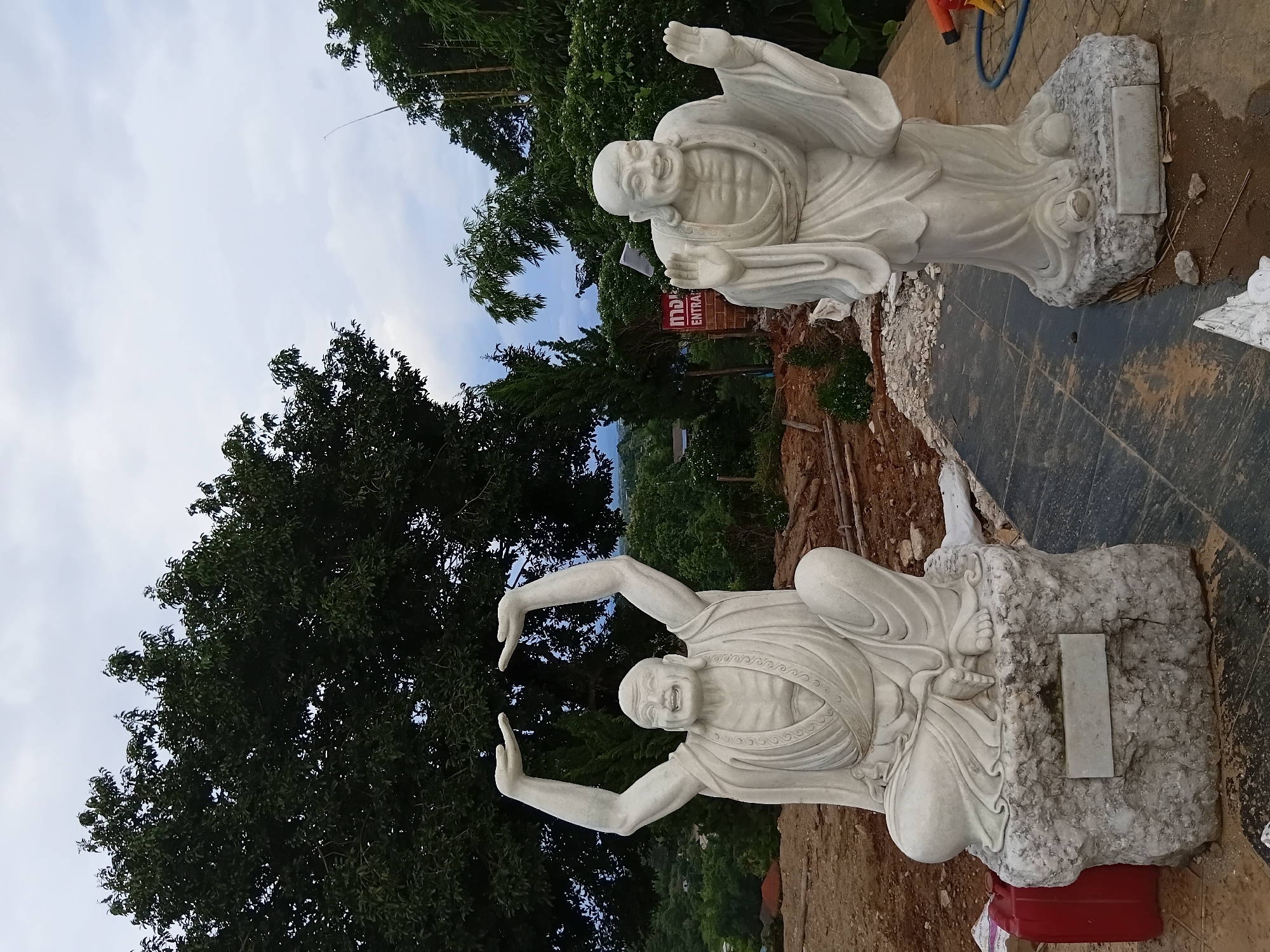 Two white marble statues of smiling Buddhist monks are positioned outdoors in an area that appears to be under construction. The statue on the left is seated with its arms raised above its head, palms open. The statue on the right stands with its arms extended outwards, palms facing forward. Both statues are intricately carved and appear to be made of polished marble. In the background, lush green trees and vegetation are visible, suggesting a natural or park-like setting. A red sign with Thai script and the word "ENTRANCE" is visible on the right side of the image. The ground is a mix of dirt, gravel, and paving stones. The sky is overcast with clouds. The overall scene depicts outdoor sculptures in a serene, yet unfinished, environment.