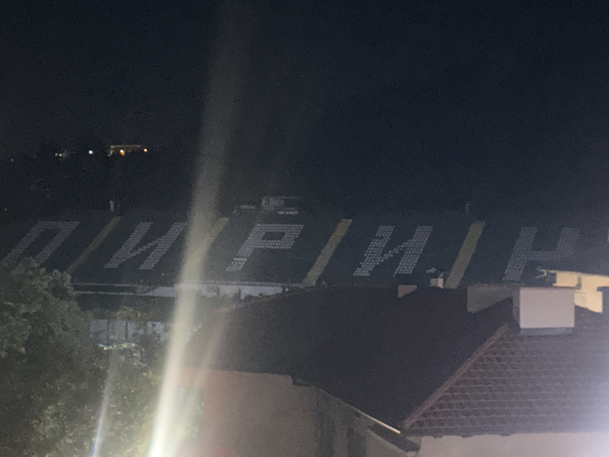 A nighttime, high-angle view captures a stadium with "ПИРИН" (Pirin) displayed in large white Cyrillic letters on its stands, interspersed with vertical yellow stripes. The stands appear dark otherwise, suggesting they are not actively lit. In the foreground, dark rooftops with tiled patterns and dense green tree foliage partially obscure the lower section of the stadium. A prominent light source in the lower-left creates a vertical lens flare across the image. On the right, a white, rectangular structure (possibly a chimney or vent) is visible on a rooftop. The background consists of a dark night sky with faint, distant lights on the horizon. The scene is located in Blagoevgrad, Bulgaria, likely depicting the Hristo Botev Stadium, home of PFC Pirin Blagoevgrad.