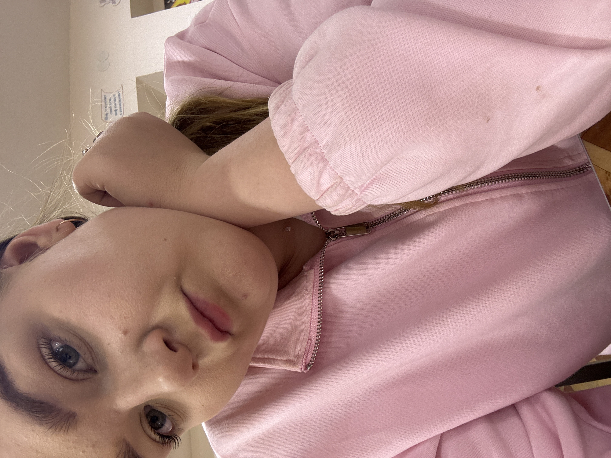 A close-up shot focuses on a fair-skinned woman wearing a pink zip-up sweatshirt. The woman's face is angled towards the viewer, with her left arm bent and her hand resting near her face. Her hair is blonde, visible beneath her arm. The zipper of the sweatshirt is silver. The background shows a portion of a wall and what appears to be furniture. There is no indication of the location being Chaikovsky, Russia.