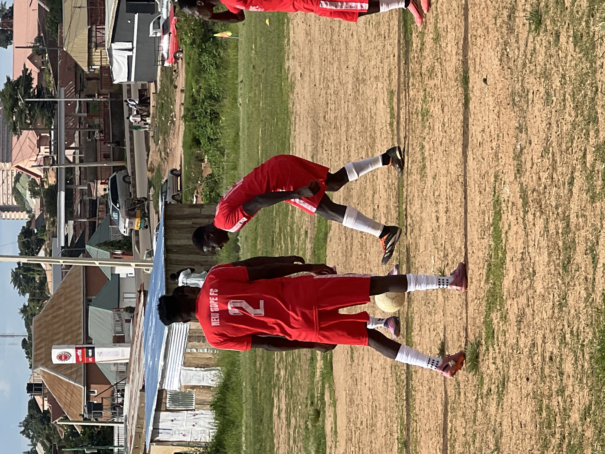 The image depicts an outdoor scene in a city, possibly Jos, Nigeria, characterized by a dusty, dry field with patches of dry grass. Several young men, appearing to be soccer players, are present. Two prominent figures in the foreground are wearing matching red soccer jerseys and shorts, with white socks pulled up to their knees. One player, facing away from the camera, has the number '2' prominently displayed on his back, along with the text 'NEW HOPE FC'. He is standing near a soccer ball. Another player, facing to his left, is in motion, with his right leg bent and his left leg extended. Other players are visible in the background, also in red uniforms.
The environment includes elements of urban development in the background: buildings with corrugated iron roofs, some with a reddish-brown hue, and others with grey or blue sheeting. A large, prominent sign with red and white lettering, likely advertising a business, stands near some buildings. The presence of cars, including a tricycle (Keke Napep), suggests a busy urban setting. The sky is clear and bright, indicating a sunny day. The overall impression is that of a soccer practice or a casual game taking place in a somewhat undeveloped area of a Nigerian city.