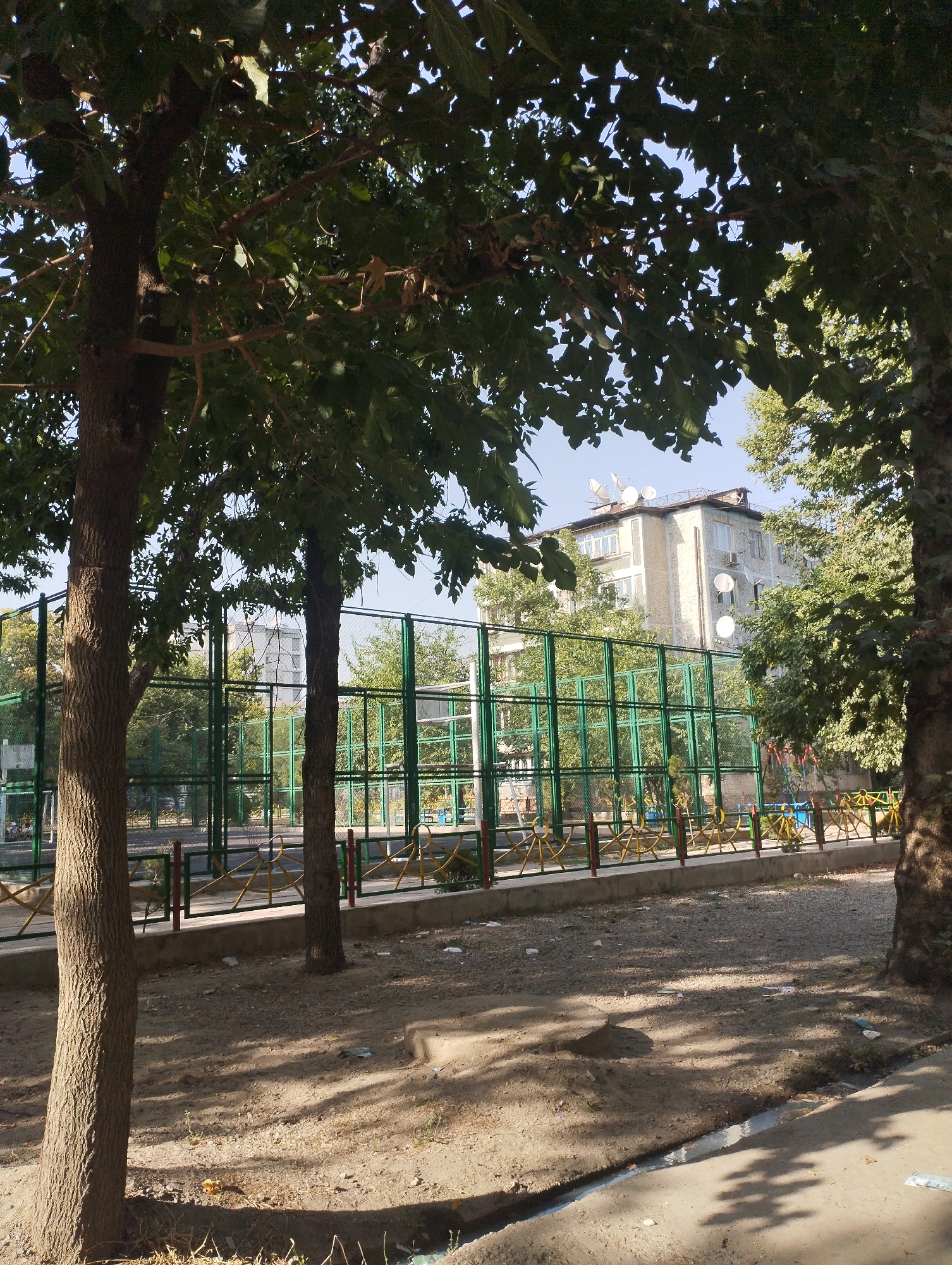 In Dushanbe, Tajikistan, this image depicts an outdoor sports court enclosed by a green metal fence. The court appears to be a basketball or tennis court, with lines visible on its surface. Beyond the fence, a multi-story building with several satellite dishes on its roof is visible. The area around the court is unpaved and appears somewhat neglected, with scattered debris. Several trees with leafy branches cast shadows on the ground, suggesting it is a sunny day. The foreground shows the ground and a drainage ditch.