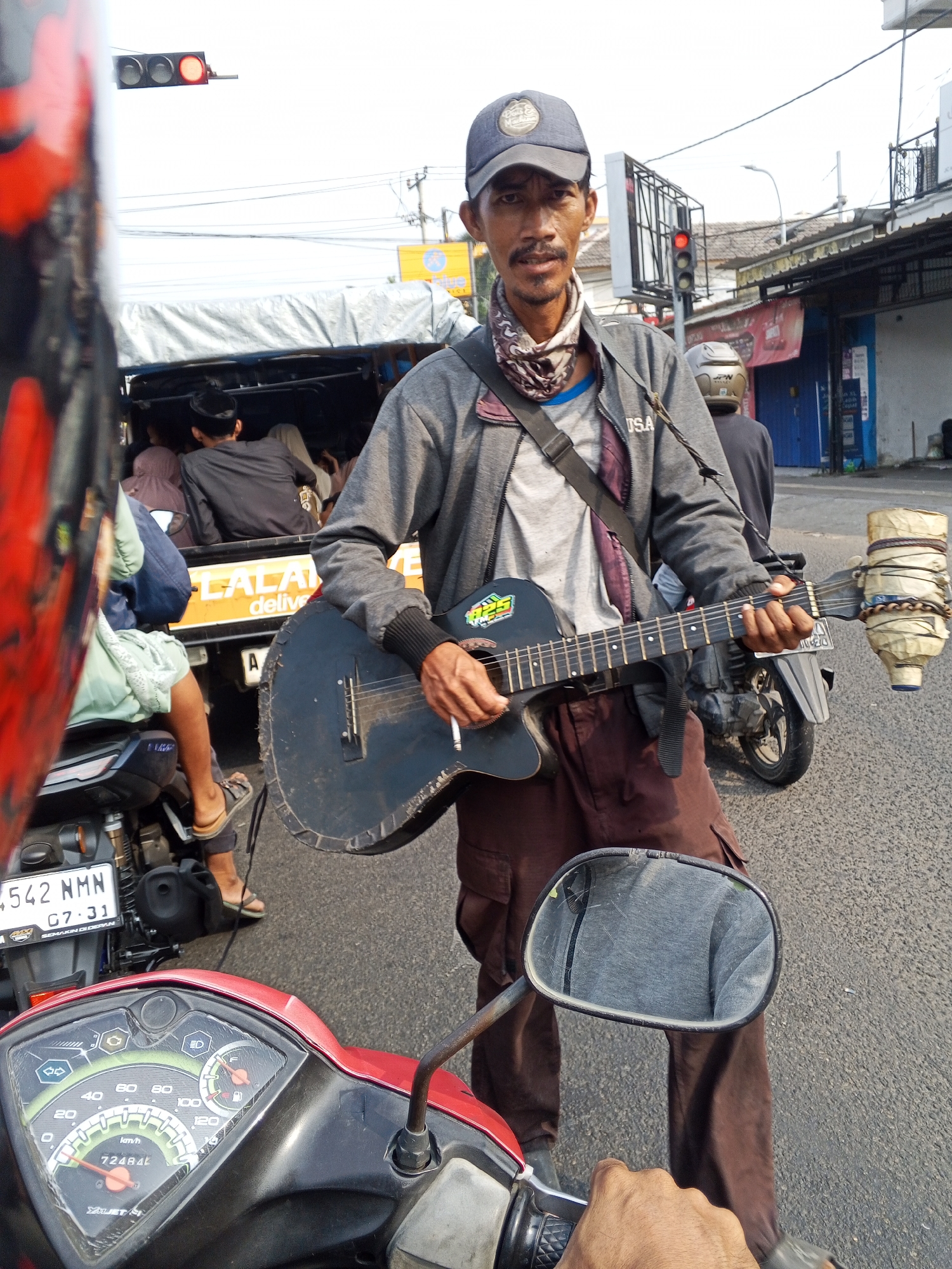 A man is playing an acoustic guitar on a street in Serang, Indonesia, while riding a red and black scooter. He is wearing a grey jacket, brown pants, a blue baseball cap, and a patterned bandana around his neck. The guitar is black and has a makeshift drum attached to its headstock. In the background, a truck is visible, carrying passengers and covered with a tarp. The street is paved and there are buildings and traffic lights in the distance. The scene appears to be a typical street view in an urban area of Indonesia.