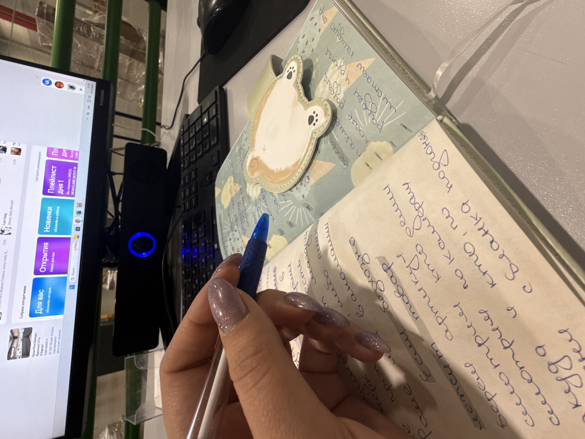 The image shows a close-up view of a hand holding a pen and writing in a notebook. The notebook is open, and there is writing in a cursive script on the pages. There is a cute, dog-shaped sticky note attached to the top of the notebook. Next to the notebook is a computer monitor displaying a webpage in what appears to be Russian, with various clickable options. A keyboard and computer mouse are also visible. The scene appears to be an indoor setting, with the computer and notebook placed on a desk. The precise location in the city of Lupanovo, Russia, cannot be determined from the image alone.