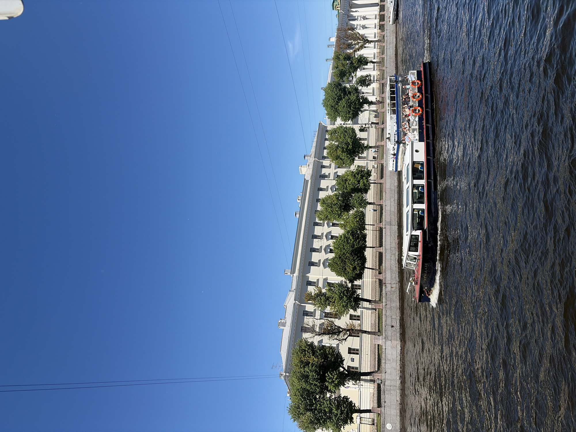 The image depicts a sunny day with a clear blue sky over a canal in Saint Petersburg, Russia. Two tour boats are navigating the dark, rippling water. The boat in the foreground, a white and blue vessel named "ЭРИДАН" (ERIDAN), is moving from left to right, creating a white wake. Several passengers are visible on its open upper deck, some wearing orange life preservers. Behind it, another, larger white boat is also moving in the same direction. Along the canal bank, a light-colored, multi-story building with numerous windows stands behind a row of lush green trees and a decorative fence. The architecture suggests it is a historical building. The overall scene captures a typical summer day with tourist activity on a waterway in the city.