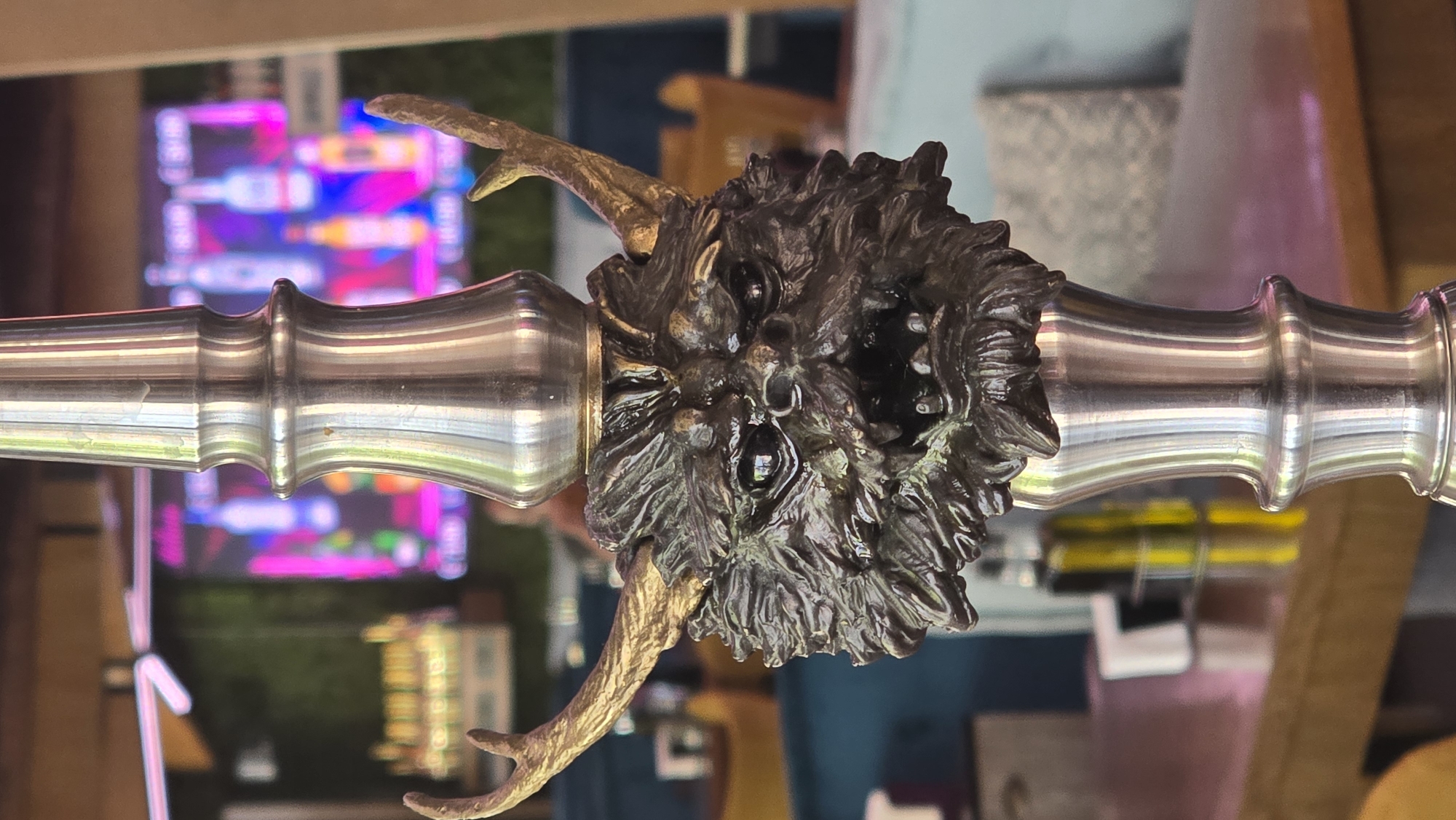 The image shows a metal object with a decorative, possibly bronze, center piece resembling a demonic face with horns. The metal pole has several bulges and indentations along its length. The background is blurred, but appears to show a colorful light display and general indoor setting. There is no information to indicate a location in Sofia, Bulgaria.