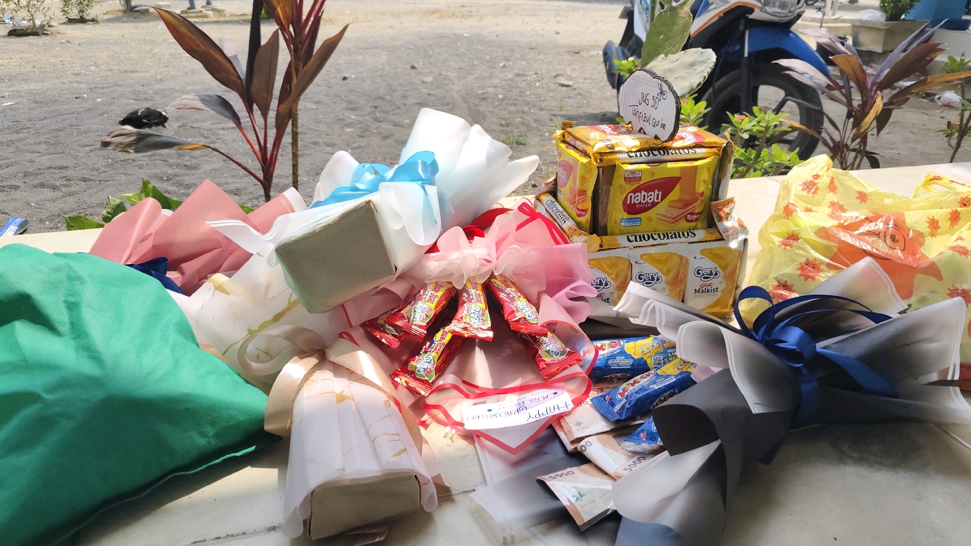 In Pangpajung, Indonesia, an outdoor scene displays a table laden with gifts. Multiple bouquets are arranged, some adorned with pink and white wrapping paper, while others feature a more subdued grey and white wrapping, tied with blue ribbon. Several snack packages are visible, including Nabati wafers, Chocolatos, and Gery Malkist. Red wrapped candies are also present. Some of the gifts appear to be money, with denominations of 5000 noted. A yellow plastic bag with an orange sun-like design sits to the right. In the background, a motorcycle is parked, and a plant with reddish-brown leaves grows nearby. The ground is covered with gravel.
