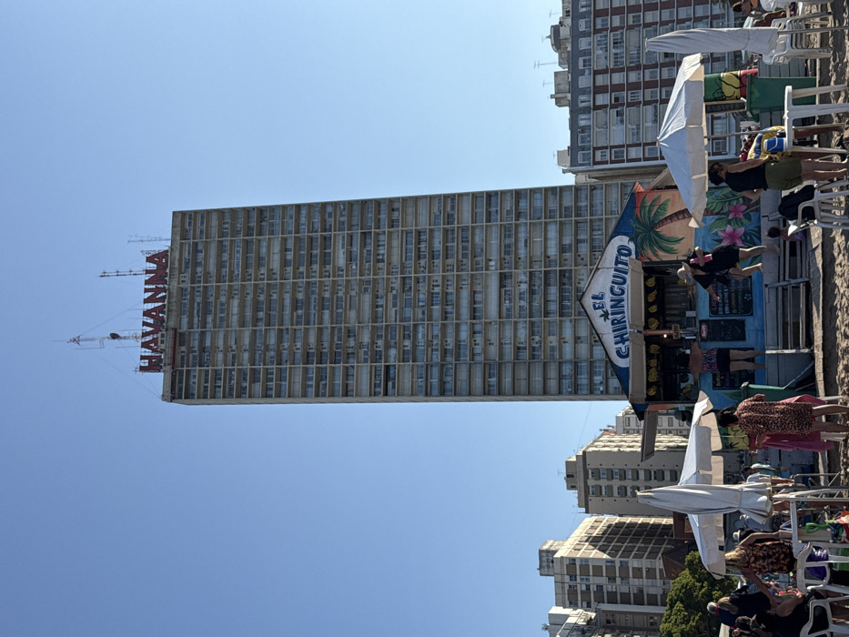 The image depicts a vibrant, sunny beach scene in Mar del Plata, Argentina, characterized by an urban backdrop and leisurely activity.
In the background, a towering multi-story building prominently displays the red letters "HAVANNA" on its roofline, serving as a distinct landmark. Other residential or hotel buildings are visible adjacent to and behind this structure, contributing to the urban coastal setting.
The foreground and midground feature a sandy beach populated by numerous individuals. People are seen relaxing in beach chairs under various umbrellas, indicating a leisure setting. A colorful beach bar, named "EL CHIRINGUITO" (Spanish for "the beach bar"), is a central element on the right side of the image. Its facade is adorned with tropical-themed paintings of palm trees and flowers. Several people are gathered near this bar, suggesting they are ordering refreshments or socializing.
The time of day is clearly bright daylight, likely mid-morning or early afternoon, with a vast, clear blue sky overhead and strong sunlight illuminating the scene. The overall atmosphere is one of relaxation and enjoyment at a bustling beach resort.