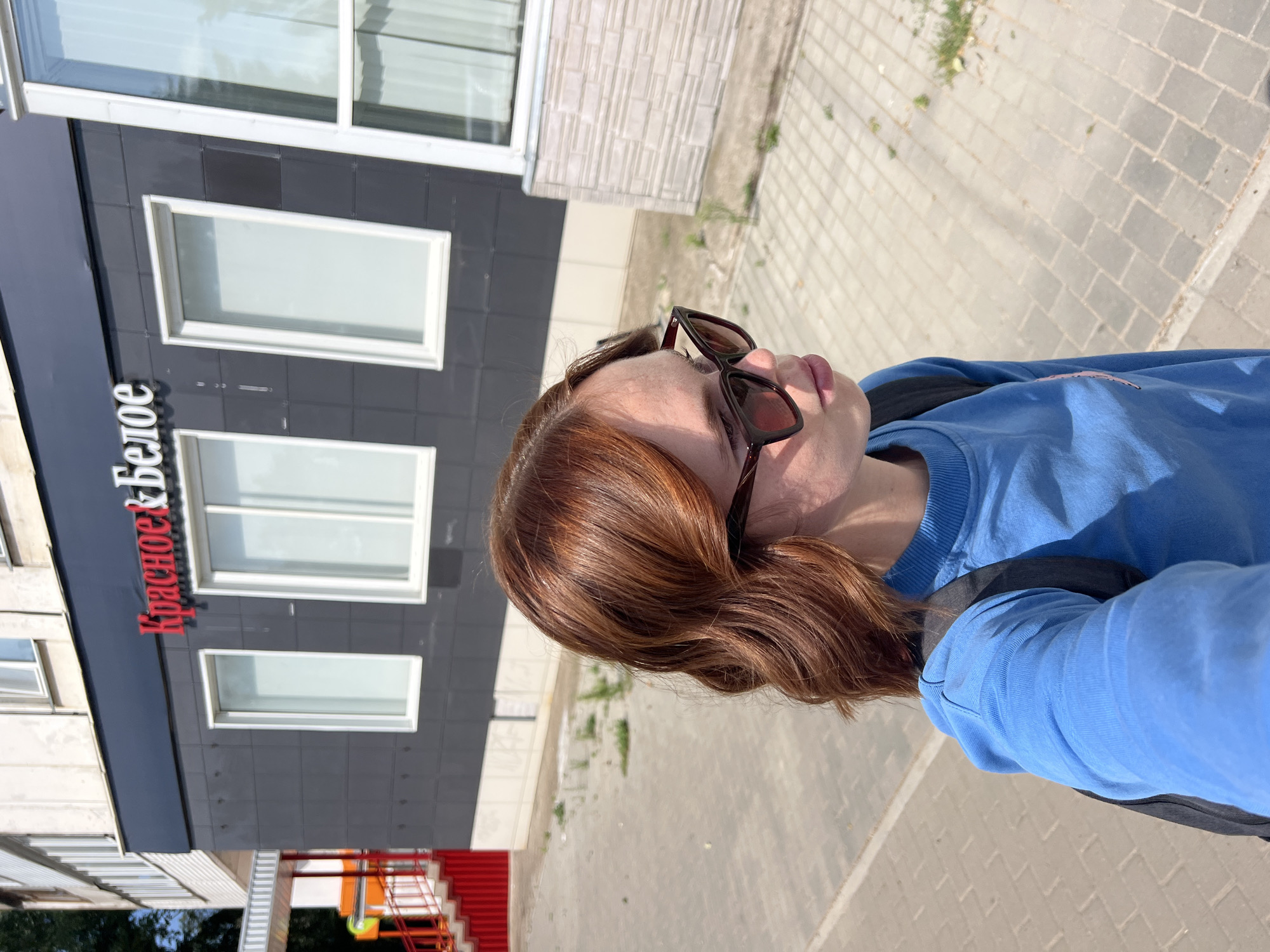 A woman with auburn hair and sunglasses stands in front of a building. The building has a dark gray facade with red and white lettering that reads "Красное & Белое," which is a Russian chain of liquor stores. The woman is wearing a blue sweatshirt and a backpack. The ground is paved with concrete tiles. In the background, a red staircase and yellow slide are visible, suggesting the presence of a playground or children's area. The overall setting appears to be an outdoor urban environment. The location is the city of Chaikovsky, Russia.