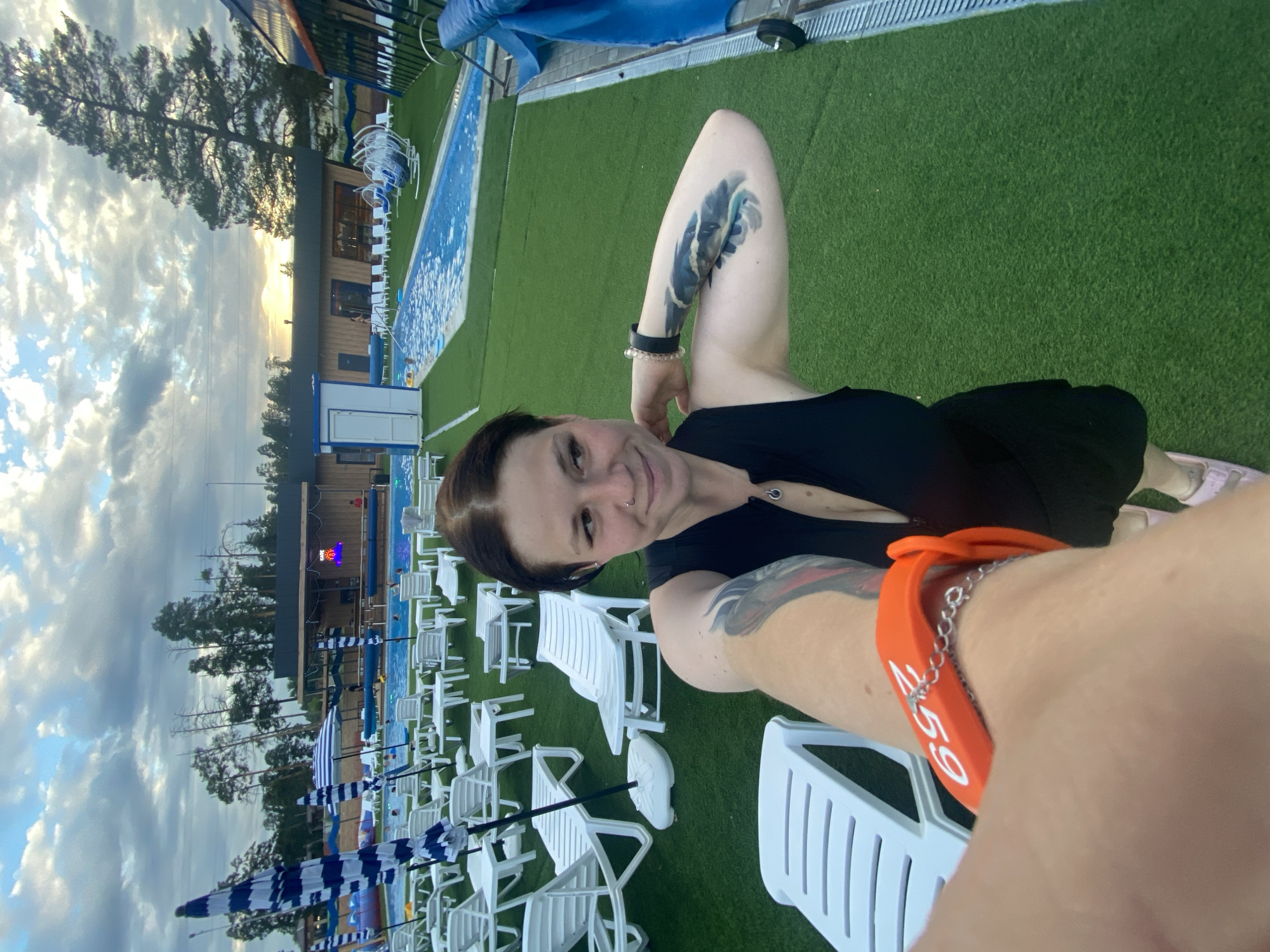 This is a selfie taken by a young woman with dark hair and tattoos on her arm. She is wearing a black dress and an orange wristband. She is standing in an outdoor area with an artificial grass surface. In the background, there are rows of white lounge chairs, blue and white striped umbrellas, and a swimming pool. There are also trees and buildings in the distance. The sky is cloudy. The location appears to be an outdoor recreation area, likely a water park or resort, potentially in Votkinsk, Russia given the reference.