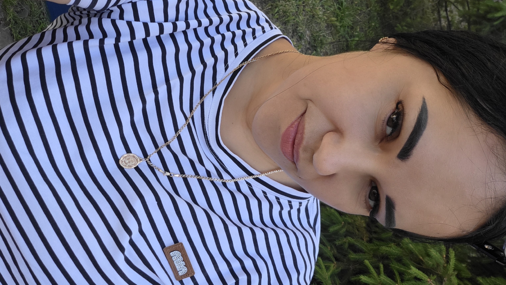 A close-up, medium shot shows a young woman with dark hair pulled back, wearing a black and white striped t-shirt and a gold necklace with a circular pendant. Her head is tilted slightly to her left, and she is looking at the camera with a subtle smile. The background consists of green foliage and grass, suggesting an outdoor setting. A small brown patch with white lettering is visible on the right side of her t-shirt. The lighting is natural, and the image appears to be taken outdoors during the day.