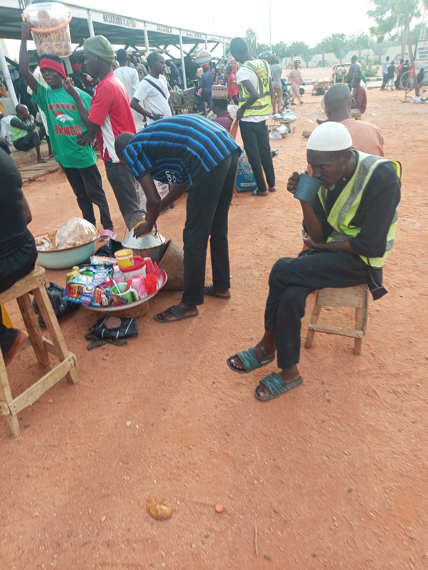 The image depicts an outdoor scene in what appears to be a bustling area of Gombe, Nigeria, based on the presence of a sign with city names including Katsina and Minna, which are cities in Nigeria. The ground is reddish-brown and appears to be dirt or gravel. Several people are visible, including vendors and passersby.
A man wearing a white cap and a yellow safety vest is sitting on a small wooden stool, holding and drinking from a blue mug. He is also wearing black clothing and sandals. Nearby, a man in a blue striped shirt is standing and seemingly attending to a food stall.
The food stall displays various food items in a tray, including sweets and snacks, next to two large bowls, possibly containing cooked food. A man is holding a container over his head. Other individuals are visible in the background, engaging in various activities.