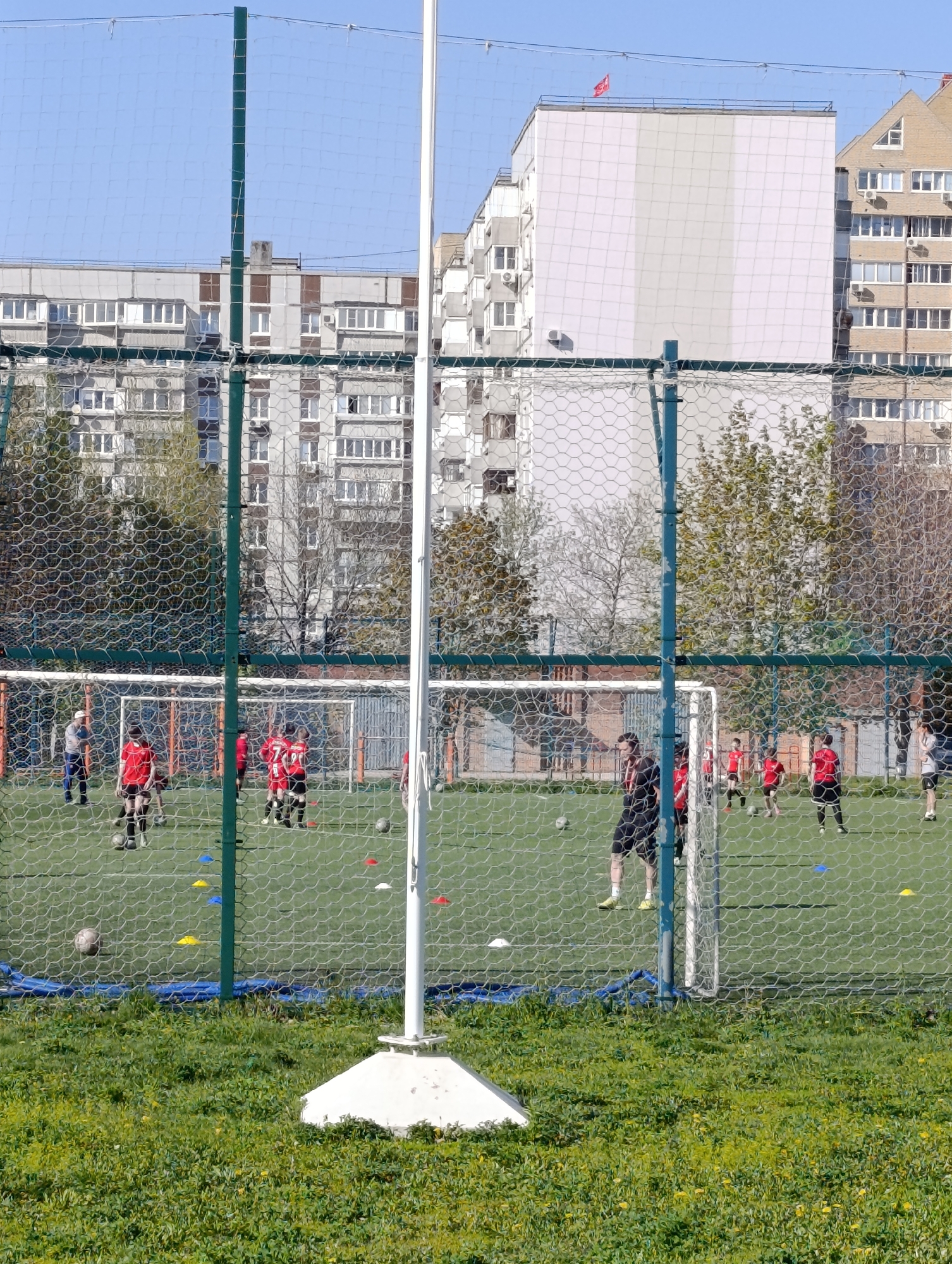 The image shows a soccer field with a green artificial turf, surrounded by a fence made of netting. In the background, there are several multi-story apartment buildings. A group of individuals wearing red jerseys and black shorts are visible on the field, seemingly participating in a practice or game. A person in black attire is observing or coaching the players. Orange and blue cones are scattered across the field, likely used as markers for drills. The foreground consists of a grassy area with yellow flowers. The overall setting suggests an urban environment. A red flag atop one of the buildings in the background is visible.
