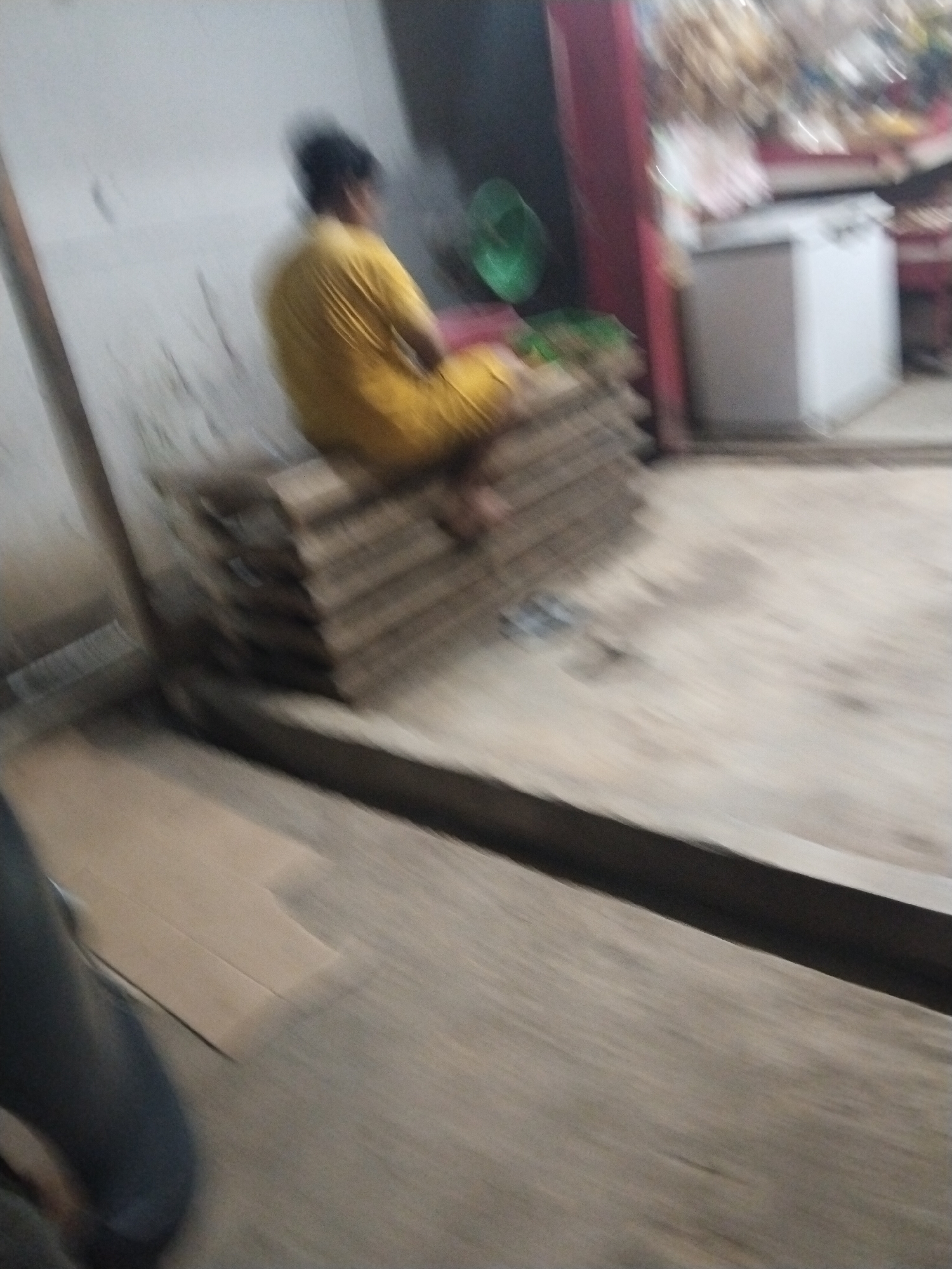 In the city of Suka Ramai, Indonesia, a person wearing a yellow garment is sitting on a stack of cardboard boxes. The scene appears to be indoors, possibly in a shop or warehouse, given the presence of what looks like shelves with items in the background and a white appliance. The lighting is dim, and the image is blurry, suggesting a low-light environment and motion. The person's face is not clearly visible due to the blurriness. The floor is made of concrete or a similar material, and a dark, horizontal line running across the image may indicate a raised edge or a shadow.