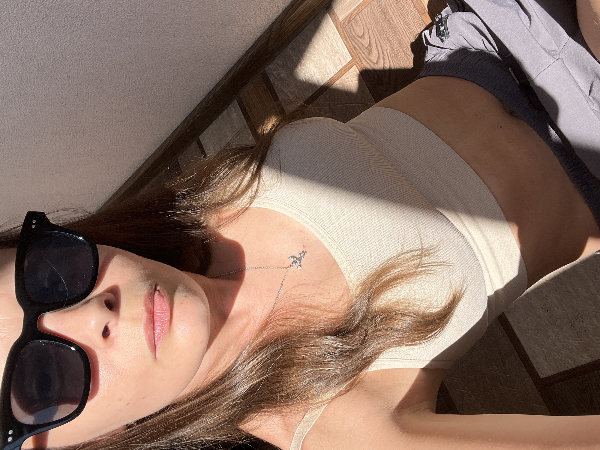 The image shows a close-up, high-angle view of a person wearing sunglasses and a light-colored tank top. The person has brown hair and is wearing a silver necklace with a small charm. They are also wearing gray shorts. The background appears to be an indoor setting with a tiled floor and a light-colored wall. Sunlight is casting shadows across the scene. No specific location in Tolyatti, Russia, can be identified from the image.