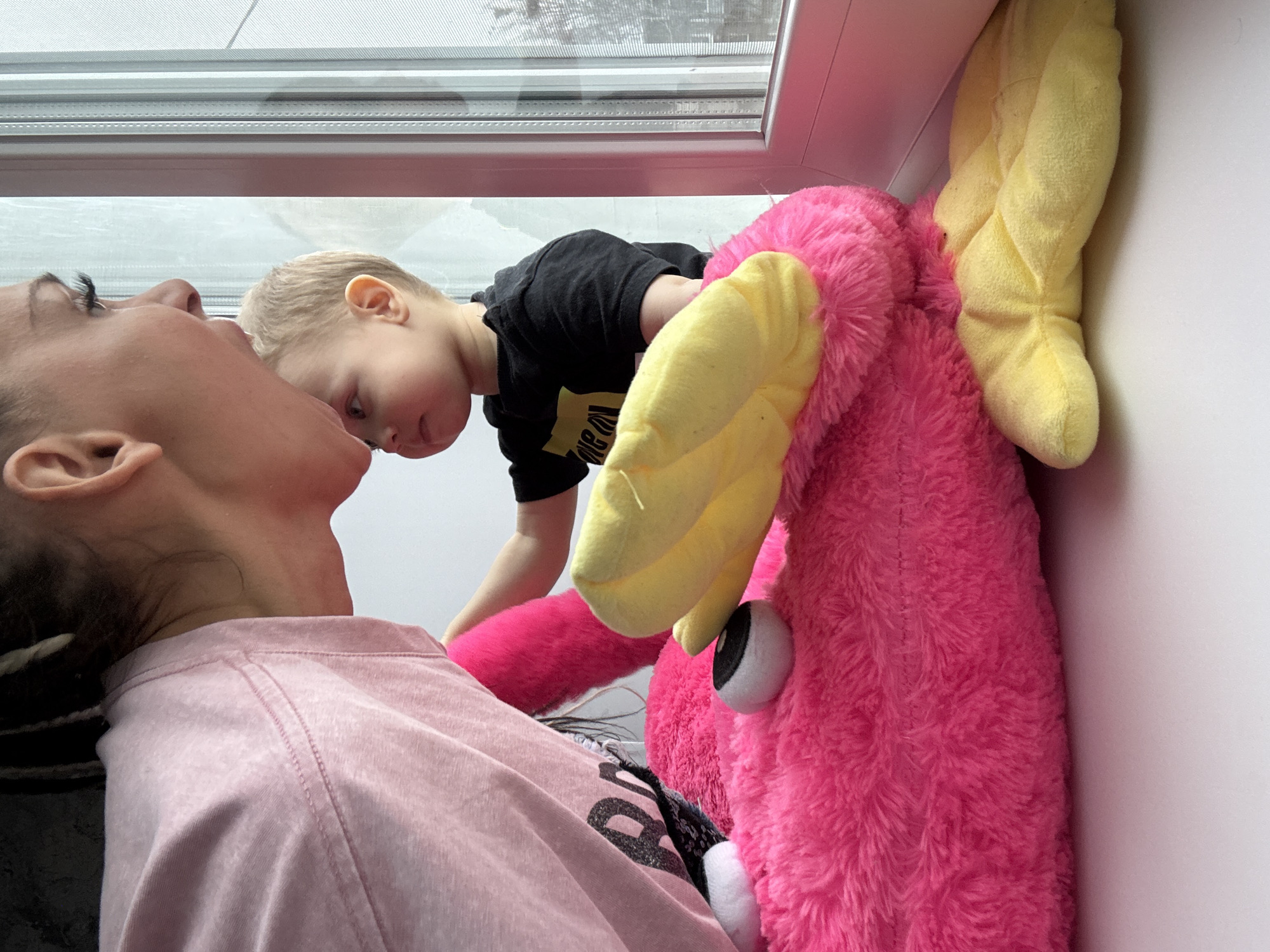 A woman is lying on her back near a window with a young child leaning over her, next to a large pink stuffed animal with yellow ears. The location is likely indoors. The exact location within Rusanovo, Russia, cannot be determined from the image alone.