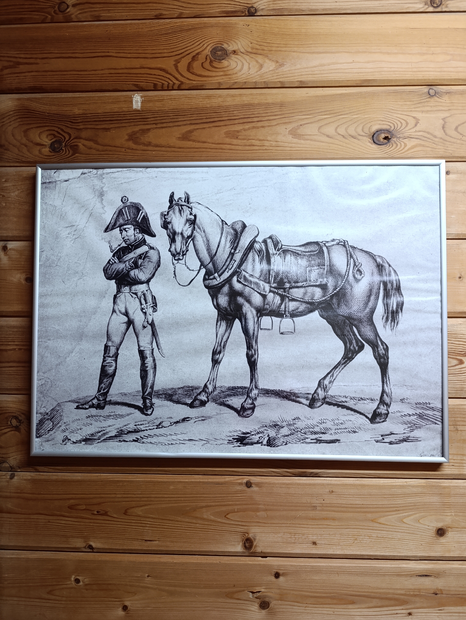 A framed black-and-white drawing depicting a man and a horse hangs on a wooden wall. The man is wearing a military uniform and a hat, and he is standing with his arms crossed. A sword hangs from his side. He appears to be smoking a pipe. The horse is walking next to the man. The wooden wall has horizontal planks with visible wood grain. The location of the scene is unknown.