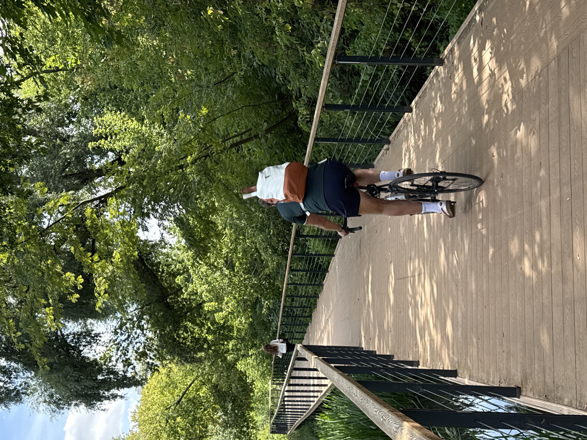 The image captures a scene on a wooden boardwalk pathway winding through a lush, green natural setting. In the foreground, a person is riding a bicycle from the perspective of someone facing away from them. The cyclist is wearing a dark green polo shirt, navy blue shorts, white socks, and brown shoes. They are carrying a white and orange backpack, with what appears to be a tennis racket extending from the top of the backpack.
Further down the boardwalk, another person is walking away from the camera, wearing a light-colored top and dark pants. They have long, dark hair. The boardwalk itself is bordered by black metal railings with wooden tops. To the left of the boardwalk, tall green reeds and calm water are visible, suggesting a marshy or wetland area. The right side of the boardwalk is lined with dense foliage and trees, creating a shaded canopy over parts of the path, casting dappled sunlight patterns. The overall environment is one of a serene park or nature trail, likely in a suburban or natural area within Moscow, Russia, given the characteristic tree types and the presence of boardwalks designed to protect natural landscapes.