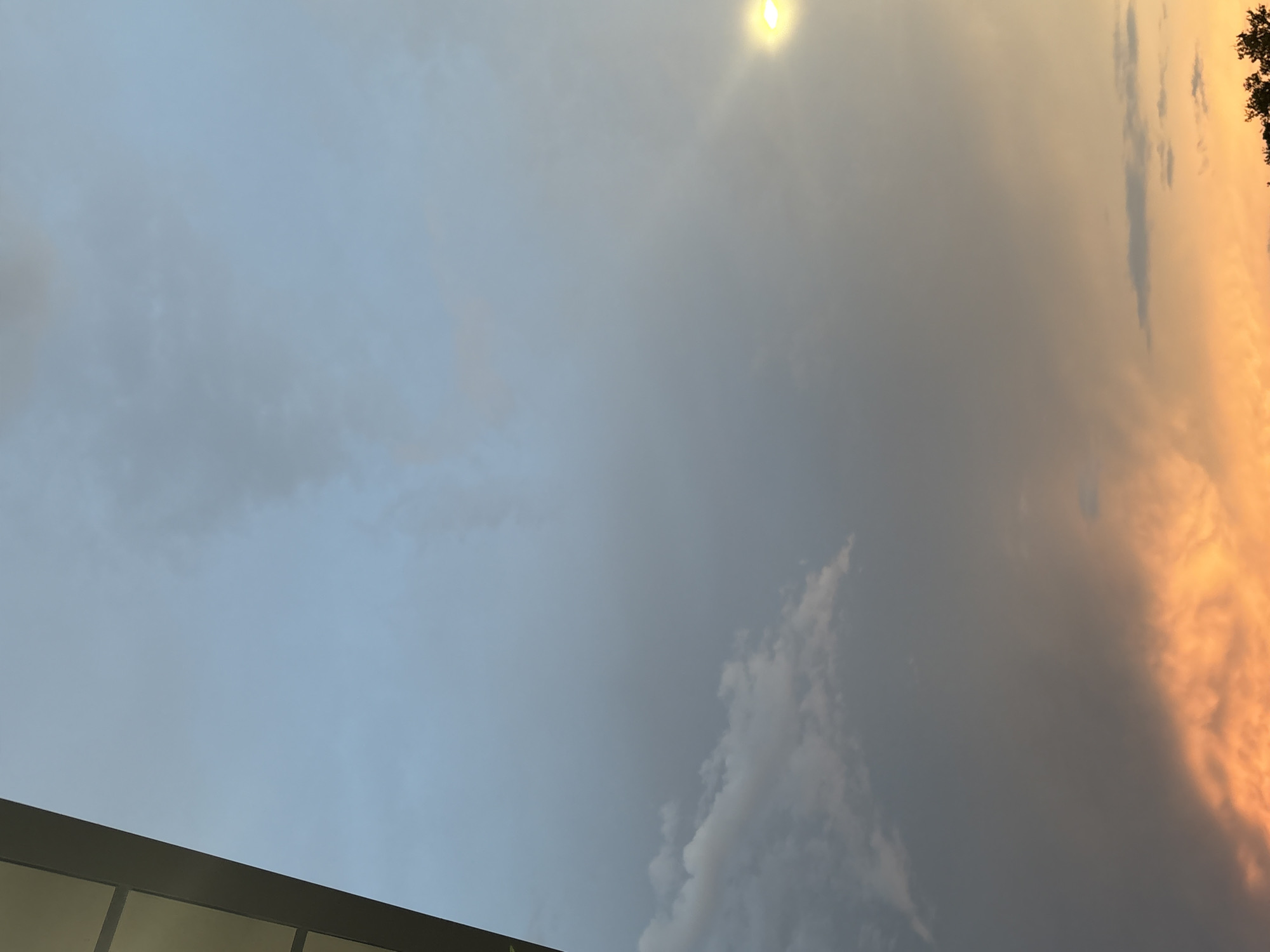 The image captures a twilight sky with dramatic cloud formations over North Port, Florida. A sliver of a building's exterior, possibly a modern structure with a dark frame and light-colored panels, occupies the upper left portion of the frame. The sky transitions from a pale blue-gray overhead to a vibrant orange and yellow hue near the horizon, indicating sunset. Several clouds are visible, some wispy and light-colored, others darker and more ominous. In the upper right, a bright light source, likely a streetlamp, casts a warm glow. The lower edge of the image reveals the silhouette of trees, suggesting a suburban or natural setting within the city. There are no people or discernible activities in the frame.