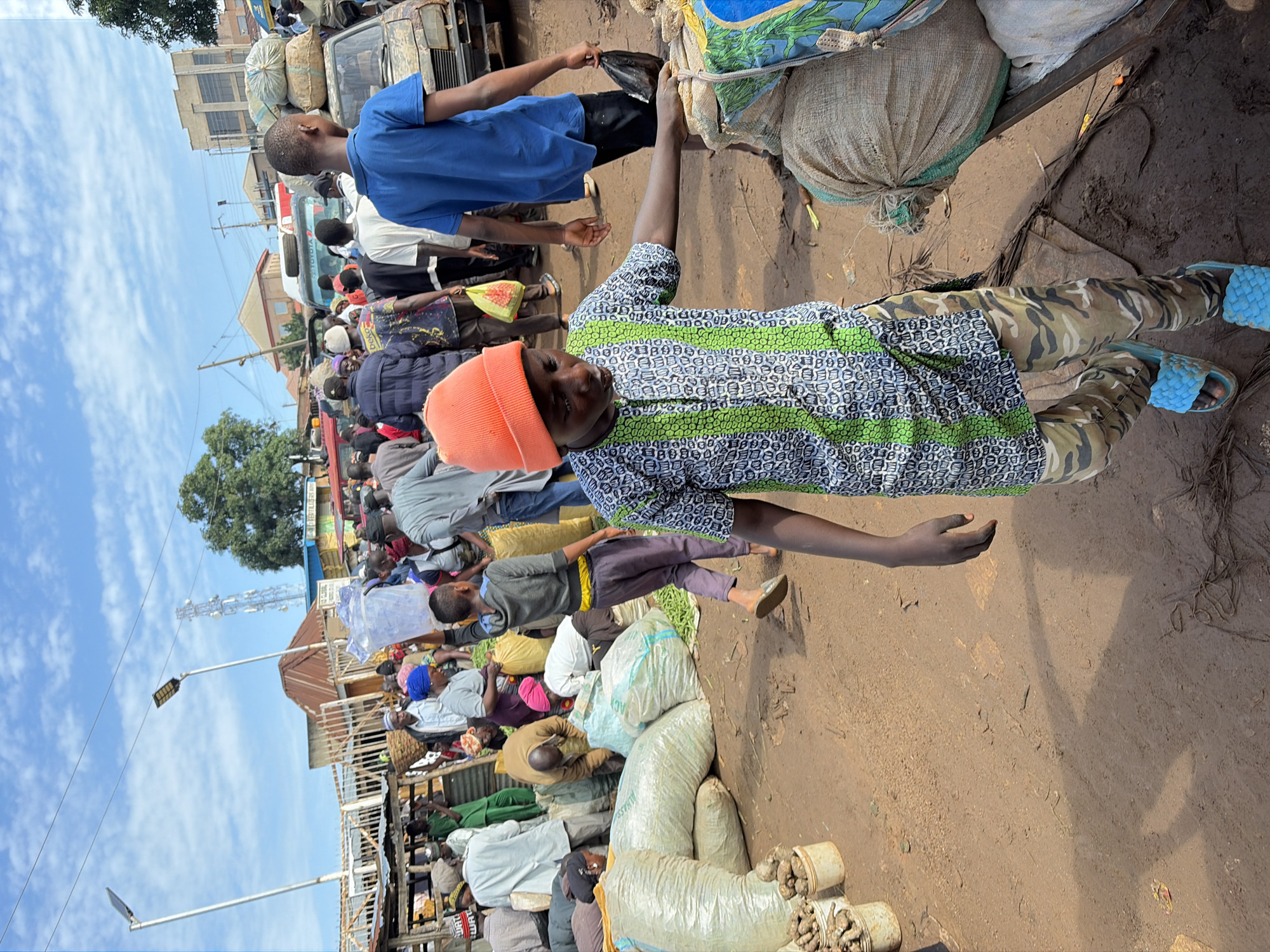 A low-angle, full shot captures a busy outdoor market scene under a bright, cloudy sky. A young boy, wearing an orange beanie, a patterned shirt with green accents, camouflage pants, and blue sandals, walks forward, reaching out towards a large burlap sack. Behind him, a crowd of people are engaged in various activities – some are carrying items, others are talking or interacting with vendors. Several large sacks of produce are visible, along with buckets containing root vegetables. A rusted vehicle, possibly a truck, is parked in the background, loaded with more sacks. The ground is dusty and uneven, suggesting a rural or informal market setting. The location is identified as Gura Top, Nigeria.