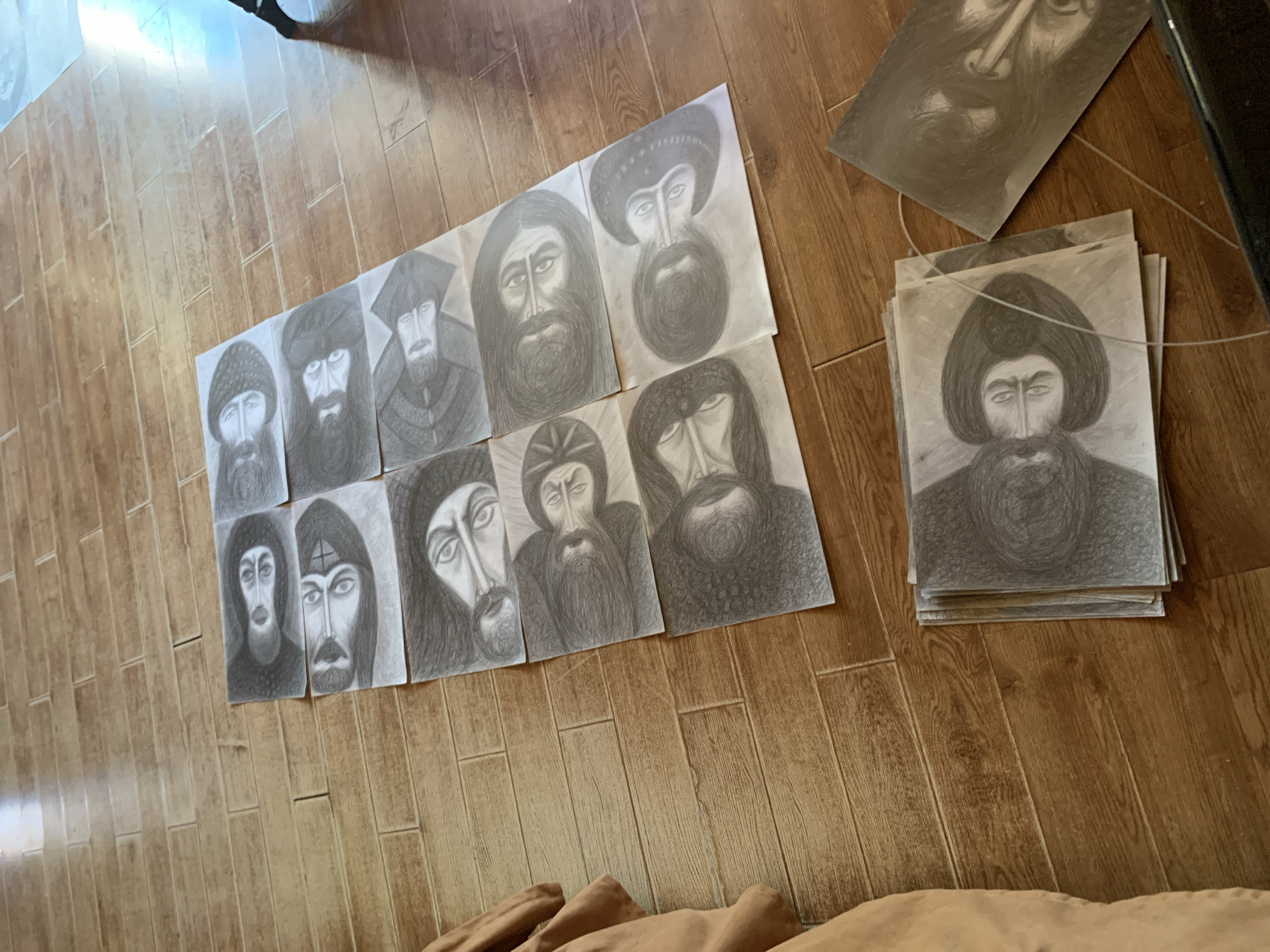 A high-angle, wide shot captures a collection of charcoal sketches of men's faces, arranged on a wooden floor. The sketches are laid out in rows, with some overlapping. Each sketch depicts a man with a beard and dark hair, rendered in varying degrees of detail. The floorboards are a medium brown wood with a visible grain pattern. To the left of the sketches, a tan fabric object, possibly clothing or a blanket, is partially visible. The lighting in the scene is bright, casting a warm glow on the wooden floor and creating subtle shadows. The background is out of focus, showing a hint of a bright light source.