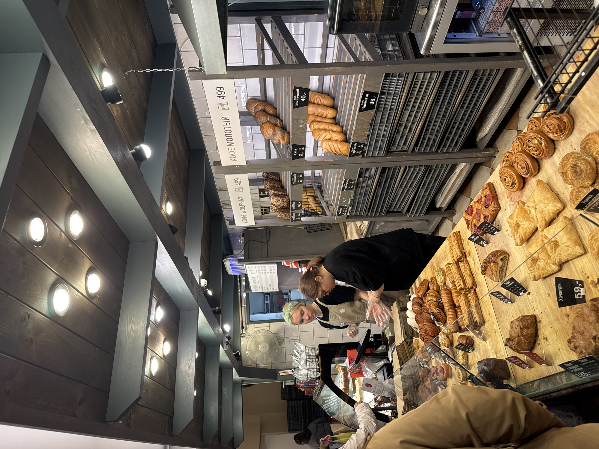 Here is a detailed description of the image:
**Scene Overview:**
The image depicts the interior of a bakery or pastry shop. The location is unidentified. The perspective is from eye-level, looking at a display counter filled with various baked goods.
**People:**
There are two individuals behind the counter, presumably bakery staff, both female. One is wearing a black shirt and the other a light-colored apron and a green headband. They appear to be handling the baked goods or assisting customers. There is also a person in the foreground, partially visible with a brown jacket.
**Objects:**
* **Baked Goods:** A wide assortment of pastries and breads are displayed, including rolls, croissants, and larger loaves. There are price tags associated with each item.
* **Display Counter:** A glass-enclosed counter presents the baked goods for purchase.
* **Shelves:** Behind the counter are shelves filled with loaves of bread and baguettes.
* **Signage:** Signs with pricing information (in Russian) are visible, including the prices for coffee ("КОФЕ").
* **Lighting:** The store has bright overhead lighting.
**Actions and Interactions:**
The bakery staff appear to be working behind the counter, possibly arranging pastries or serving a customer. A customer is present in the foreground.
**Environment:**
The bakery has a rustic yet modern aesthetic. It features a wooden ceiling with track lighting and displays. The walls appear to be tiled.