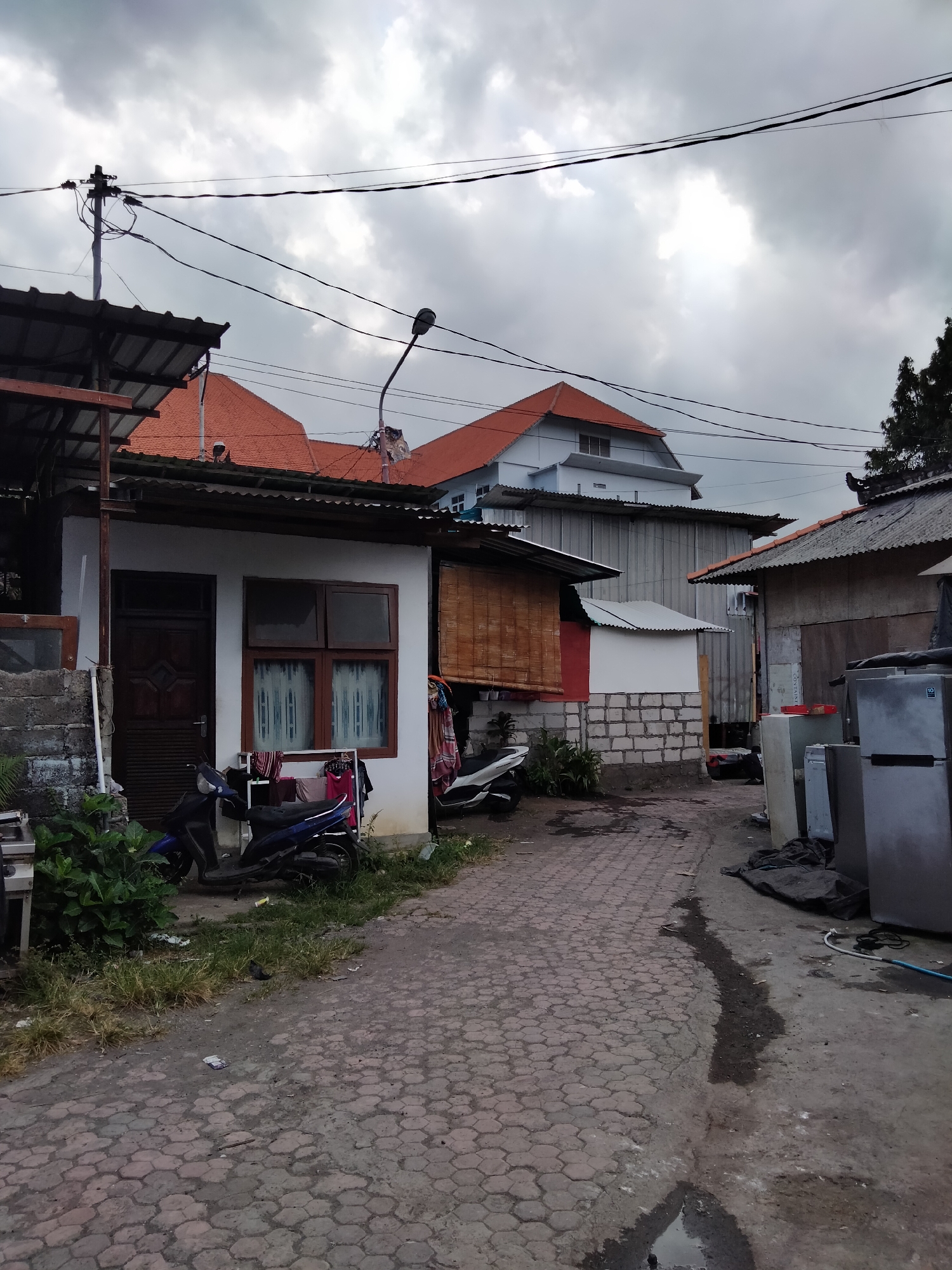 The image depicts a narrow, paved alleyway in a residential area of Denpasar, Indonesia. The alley is lined with modest, single-story houses and some larger buildings in the background. Overhead, a tangled network of power lines crisscrosses the cloudy sky.
On the left side of the alley, a small white house with a dark wooden door and two windows with brown frames is visible. Laundry is hanging outside the windows. A blue motorcycle is parked in front of the house, next to a patch of green vegetation.
Further down the alley, more houses with corrugated metal roofs and brick walls can be seen. There are several large appliances, including what appear to be refrigerators, stacked along the right side of the alley. The ground is paved with hexagonal bricks, and there are puddles of water, suggesting recent rain. The overall impression is of a working-class neighborhood with a somewhat cluttered and utilitarian appearance. The overcast sky and the presence of laundry suggest it is daytime, possibly late afternoon. No people are visible in the image.