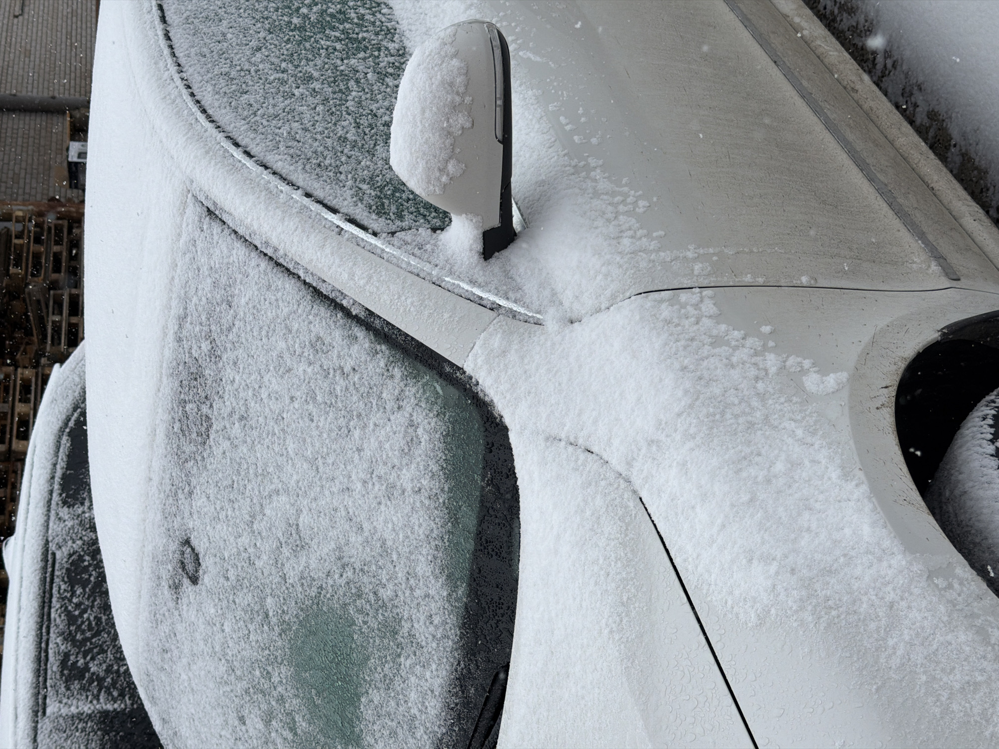 A close-up shot shows a car covered in snow, possibly in Chaikovsky, Russia. The snow is evenly distributed on the windshield, side mirror, hood, door, and the tire visible in the frame. The vehicle is a light color. The background includes the side of a building, also with some snow accumulation.